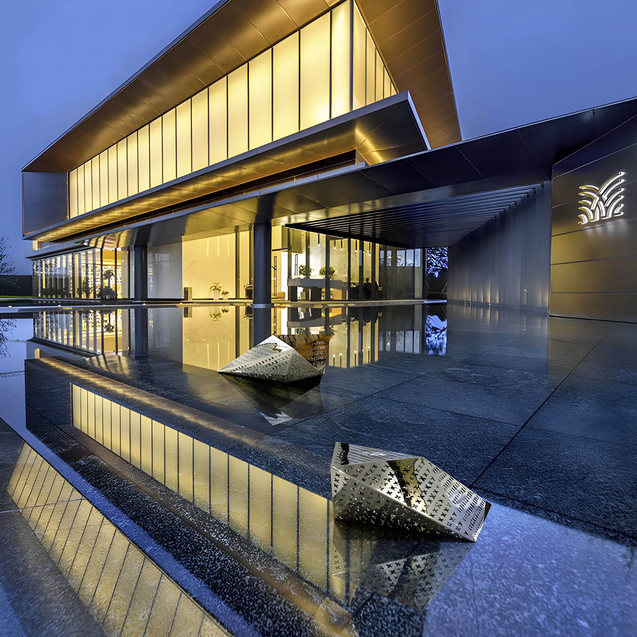 A modern building with large glass windows and warm lighting reflects on a sleek, wet surface. Two Edge Geometric Steel Outdoor Sculptures by Giant Sculptures sit in the foreground under an overcast sky, enhancing the buildings illumination.