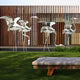 The Flamingo Stainless Steel Outdoor Sculpture by Giant Sculptures, in both 94cm and 147cm sizes, elegantly adorns a grassy lawn in front of a wooden slatted building. A stone bench completes the contemporary outdoor scene with abstract metallic bird designs.