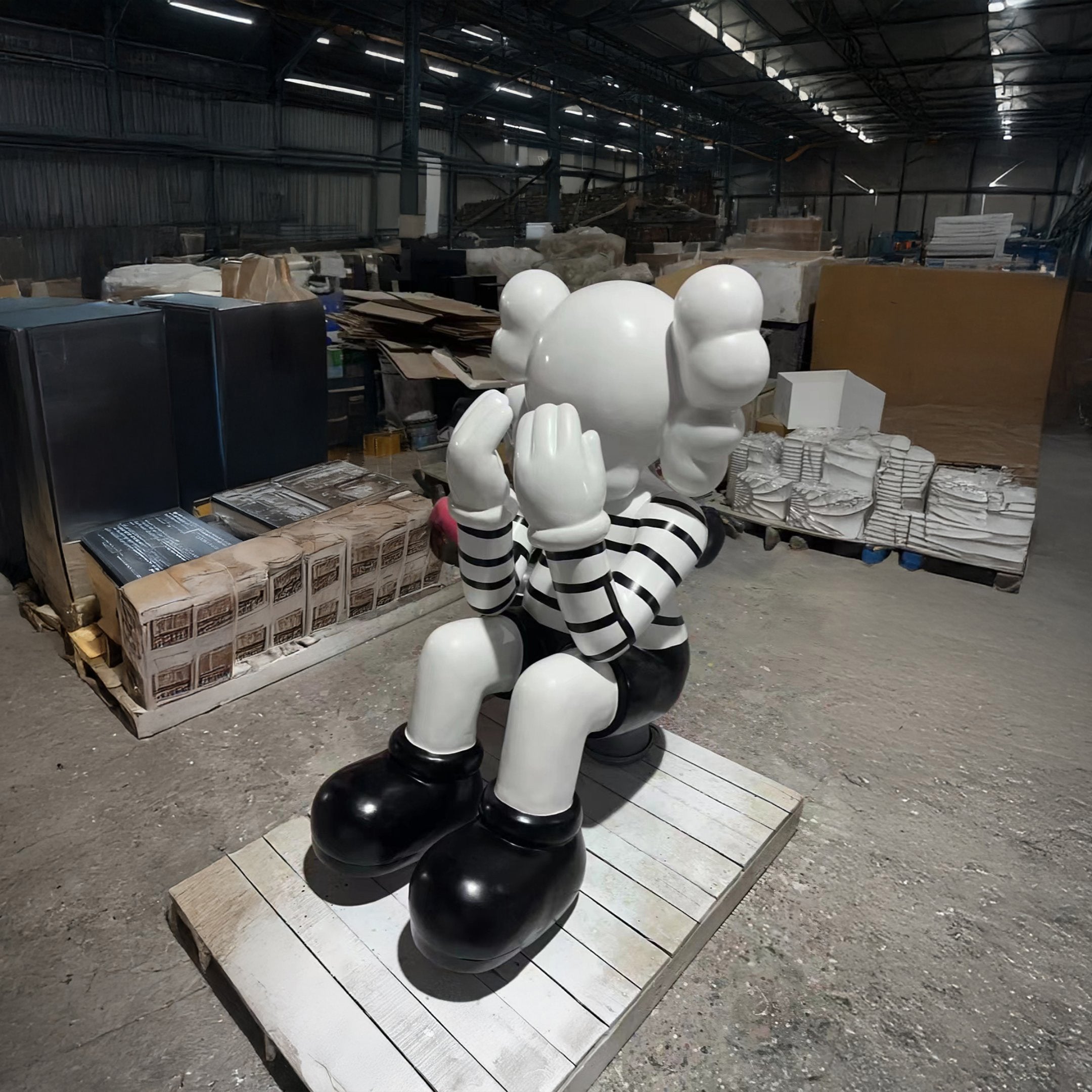 A large indoor sculpture of a seated character with a white face and round, balloon-like ears, dressed in a black-and-white striped shirt, black shorts, and large black shoes, hands covering the face in a pose of contemplation or sadness. The sculpture is positioned on a wooden pallet in an industrial setting, surrounded by warehouse items and boxes.