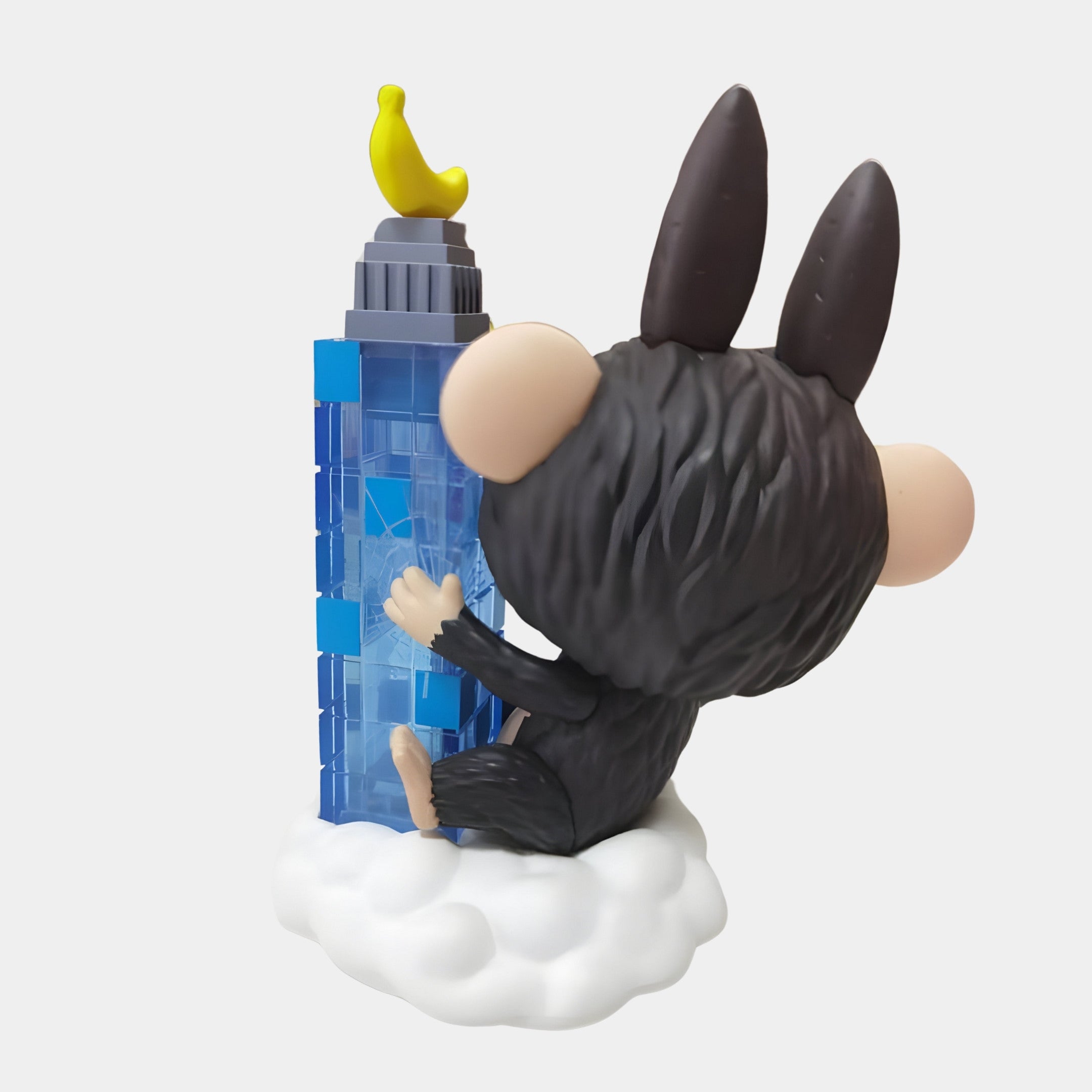 The Giant Sculptures Monsterverse Gorilla King Labubu Sculpture - 160cm features the figure with large ears sitting on a white cloud, holding a blue skyscraper topped with a yellow banana, highlighting its back and fluffy texture.