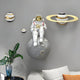 The Giant Sculptures White 3D Galaxy Astronaut On Moon Wall Art features a resin astronaut on a moon sphere, surrounded by ringed planet-like orbs. Below, a white couch with flowers and a plant accentuates its cosmic allure.