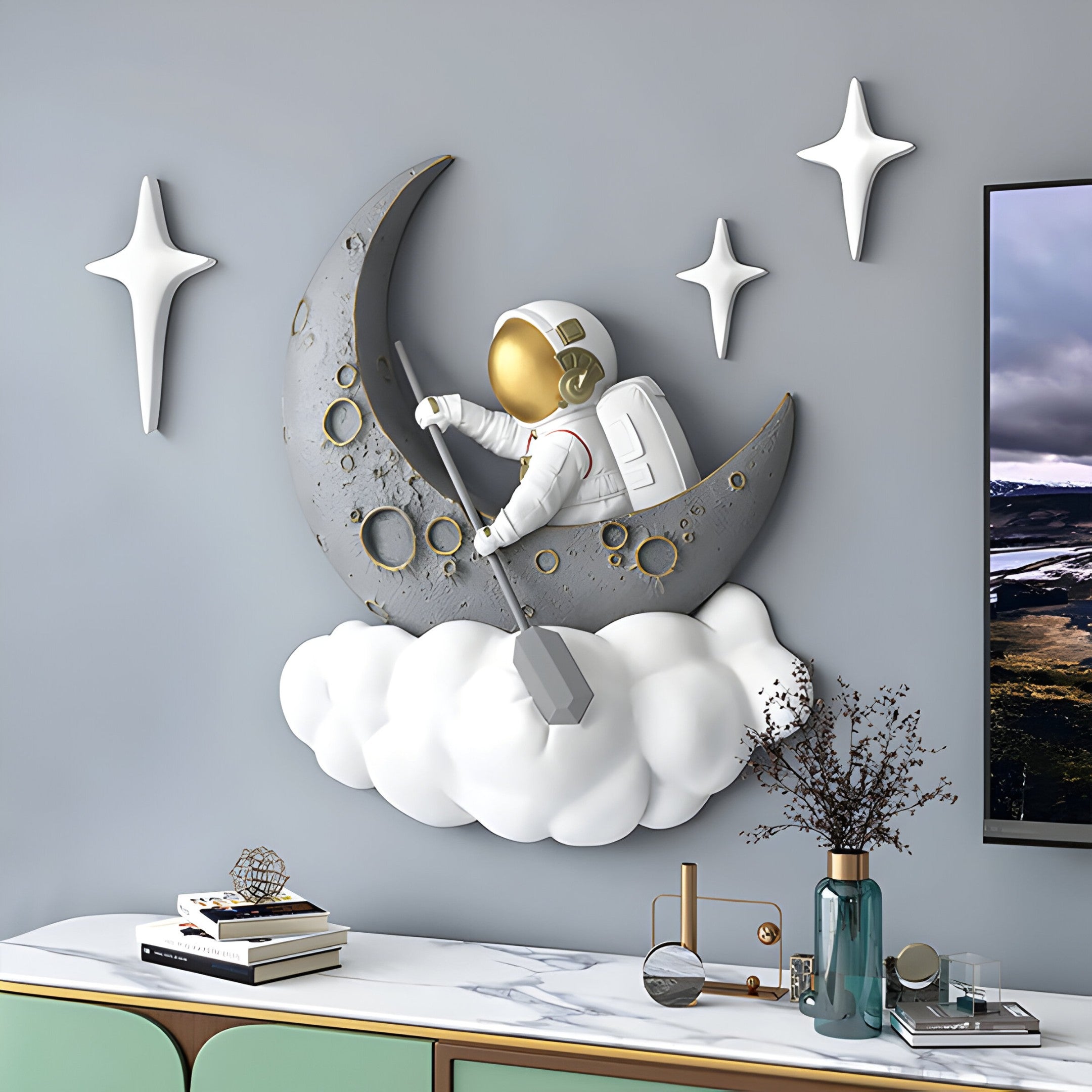 The 3D MoonSail Astronaut On Clouds Wall Art by Giant Sculptures features an astronaut in a white suit with a gold visor, rowing on a crescent moon surrounded by clouds. Three star decorations sparkle above, with items like a vase and books adorning the sideboard below.