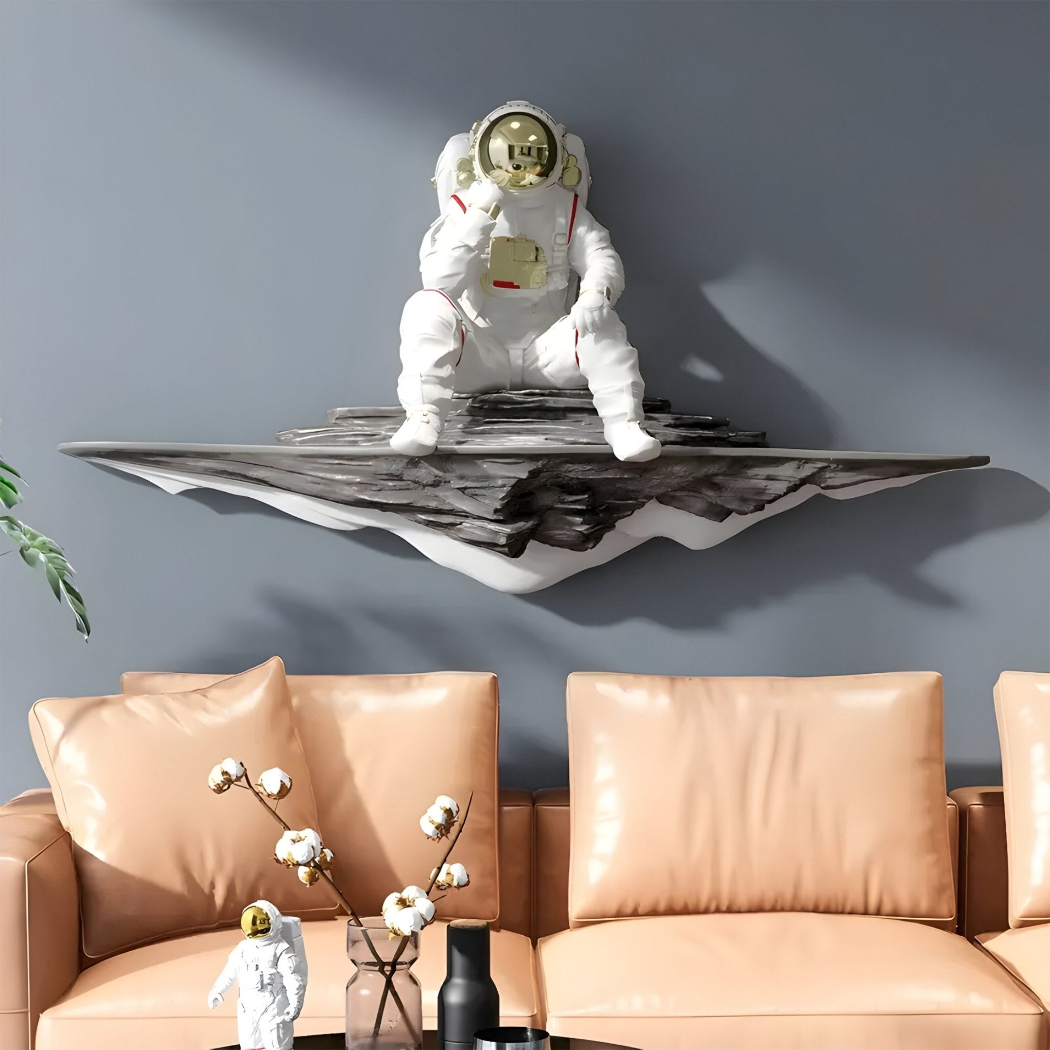 The White Astronaut Thinker On Rocky Shelf 3D Wall Art by Giant Sculptures complements a tan leather couch featuring a small astronaut statuette and black vase with white flowers, against a soft gray wall that perfectly highlights the cosmic décor.