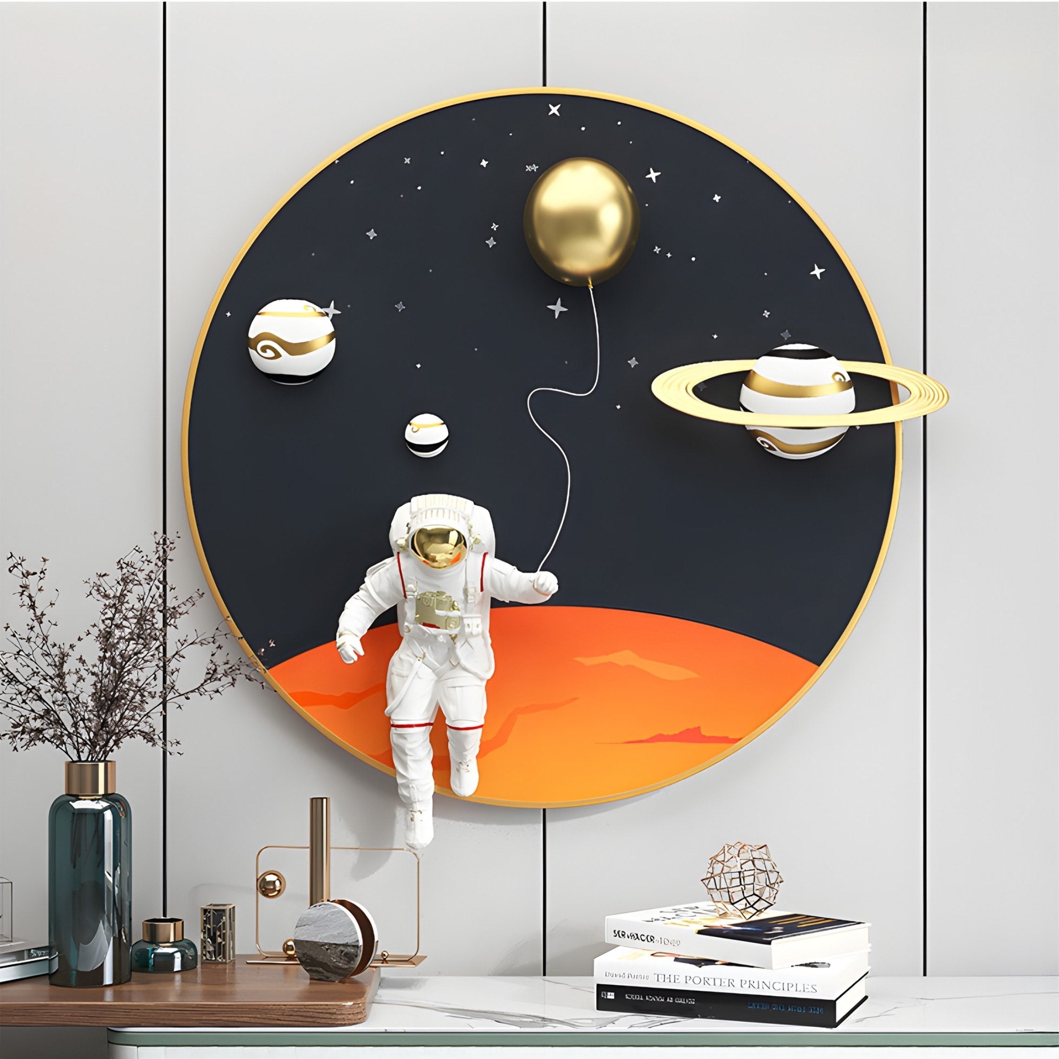 The White 3D Space Astronaut Balloon Wall Art by Giant Sculptures showcases an astronaut with a golden balloon on a red planet, surrounded by cosmic planets and stars on a circular framed black background. A vase, books, and small sculpture complete the scene below.