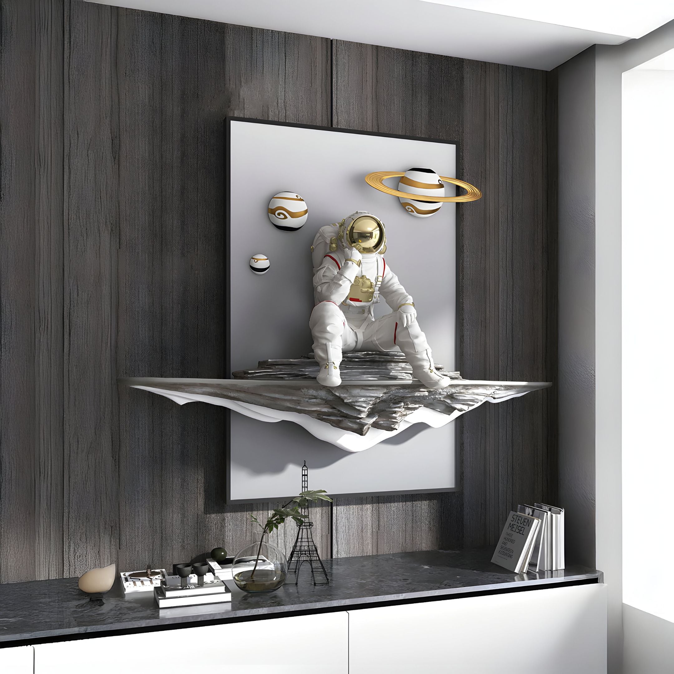 The White 3D Galaxy Astronaut Thinker Wall Art by Giant Sculptures features a resin astronaut seated on abstract terrain with planets in the backdrop. Its mounted above a modern counter with decor, adding cosmic introspection to the contemporary rooms ambiance.