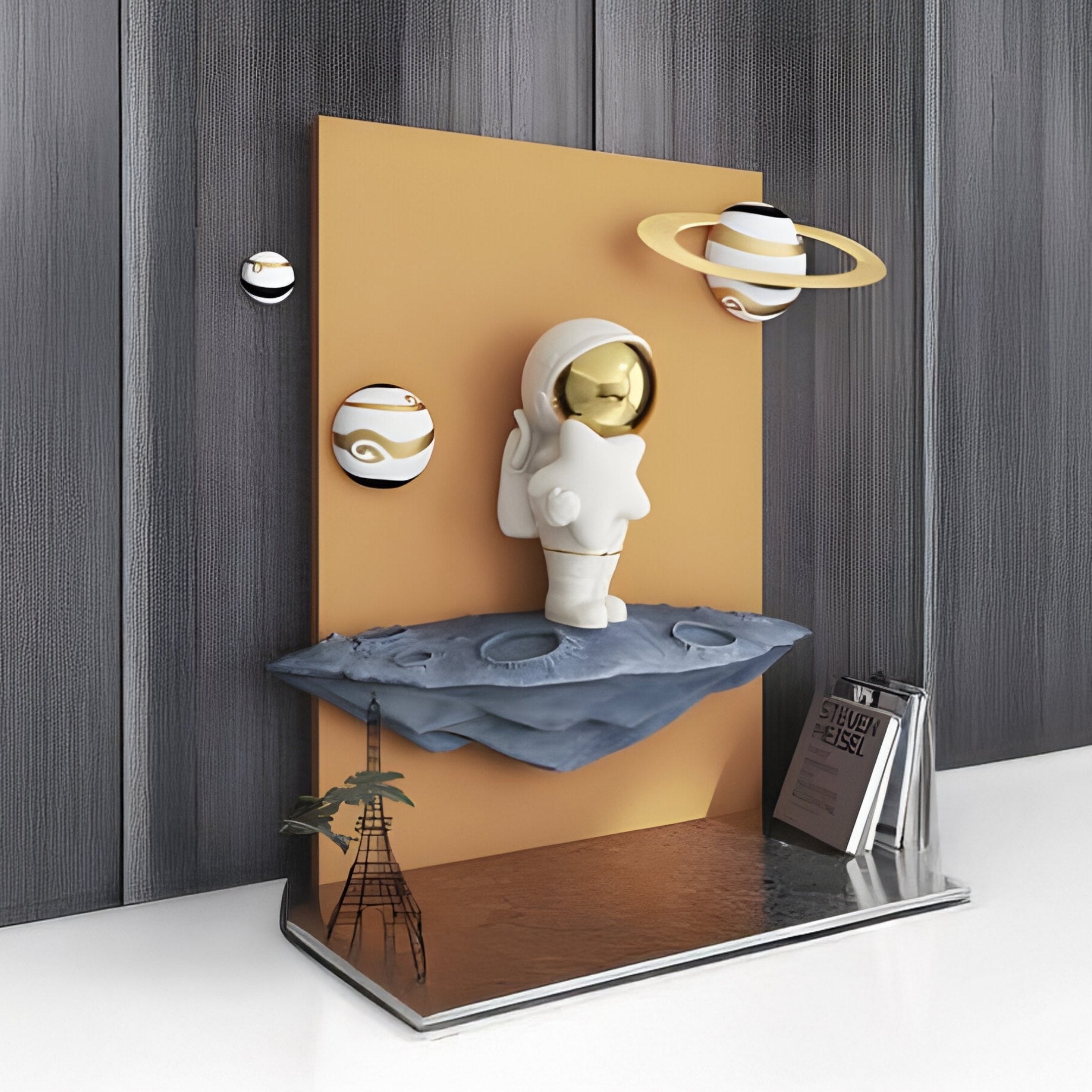 The White 3D Stardust Astronaut Wall Art by Giant Sculptures features a figurine on a gray surface with a yellow background, floating orb-like planets, and includes a book and tower model for an imaginative, futuristic look.