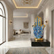 The Golden Hamsa Hand Protection Sculpture - 150cm by Giant Sculptures, featuring blue and gold accents and a central eye, stands on a black pedestal in a luxurious marble hallway with modern lighting and arched windows.