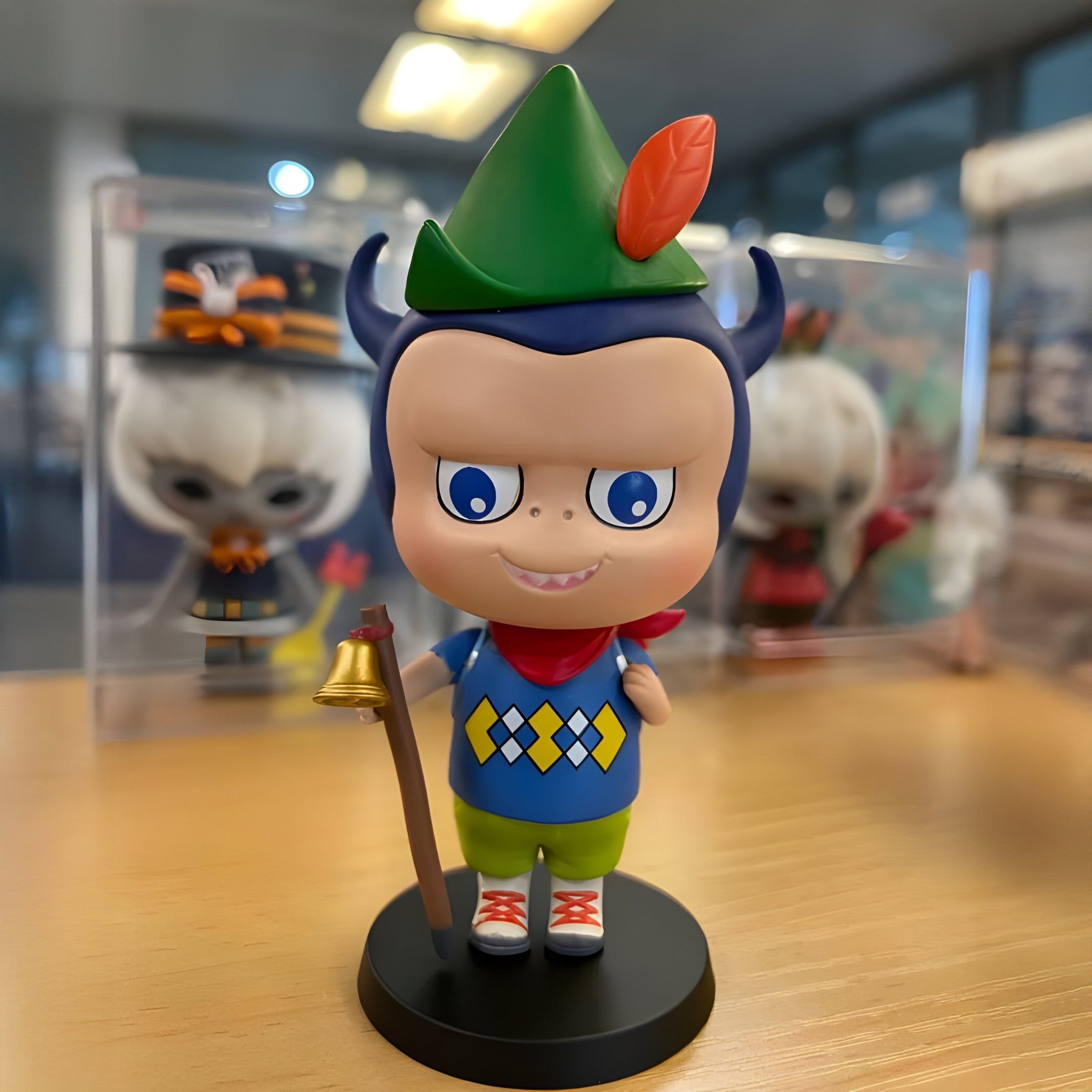 The Camp Life Forest Scout Labubu Sculpture (100cm) by Giant Sculptures features blue horns, a green hat with an orange feather, big blue eyes, and a mischievous smile. It stands on wood holding a staff, with blurred collectibles in the background.