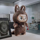 The Giant Sculptures The Monster I Found You Zimomo Labubu Sculpture (100cm) stands on a desk, its bunny ears and mischievous grin brightening a design-led workspace as it holds a small red object among office supplies.