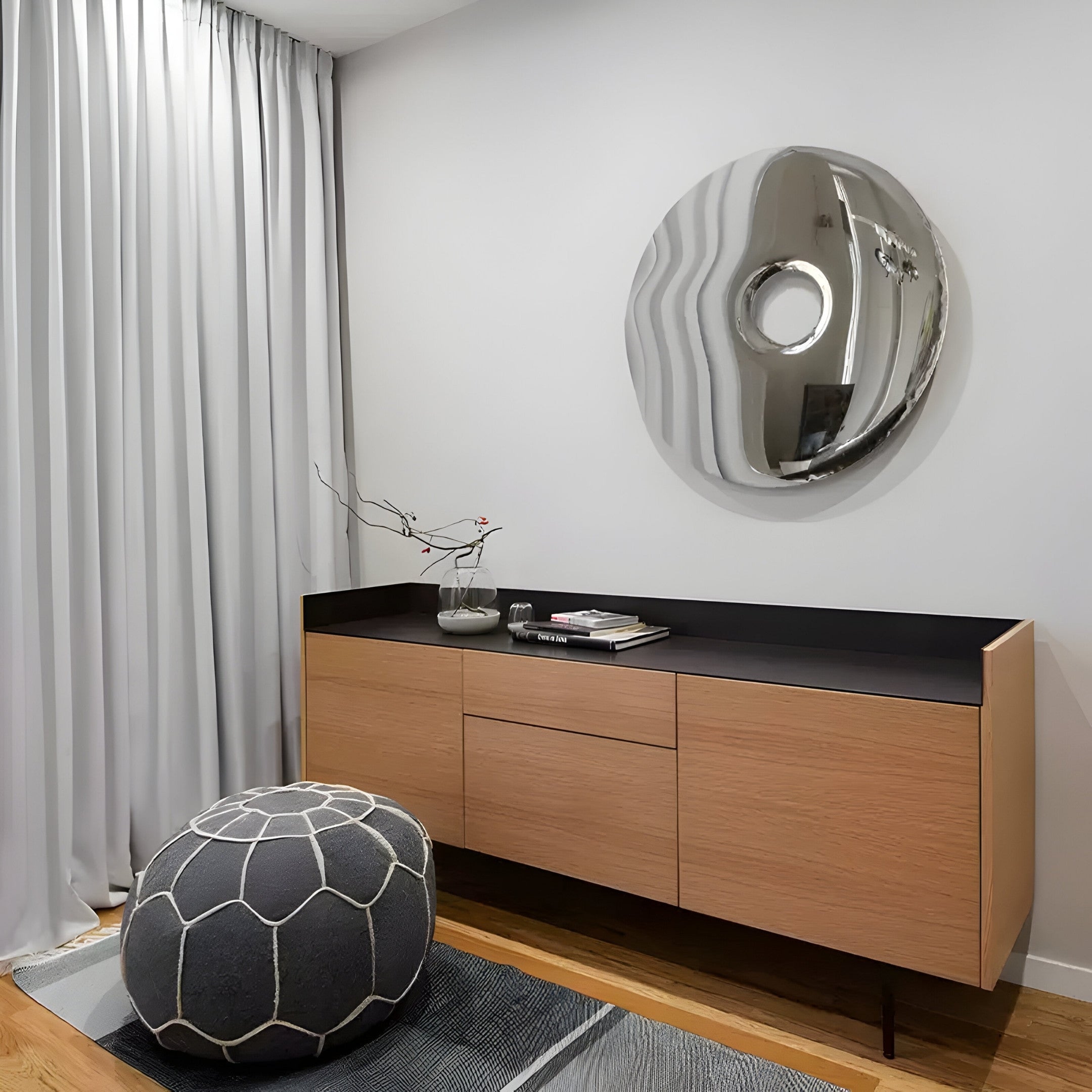 In a modern living room, a wooden credenza with a black top holds decor and magazines. The Auro Chrome Round Gloss Metal 3D Wall Art by Giant Sculptures hangs above it. A patterned ottoman rests on a striped rug, framed by gray curtains.