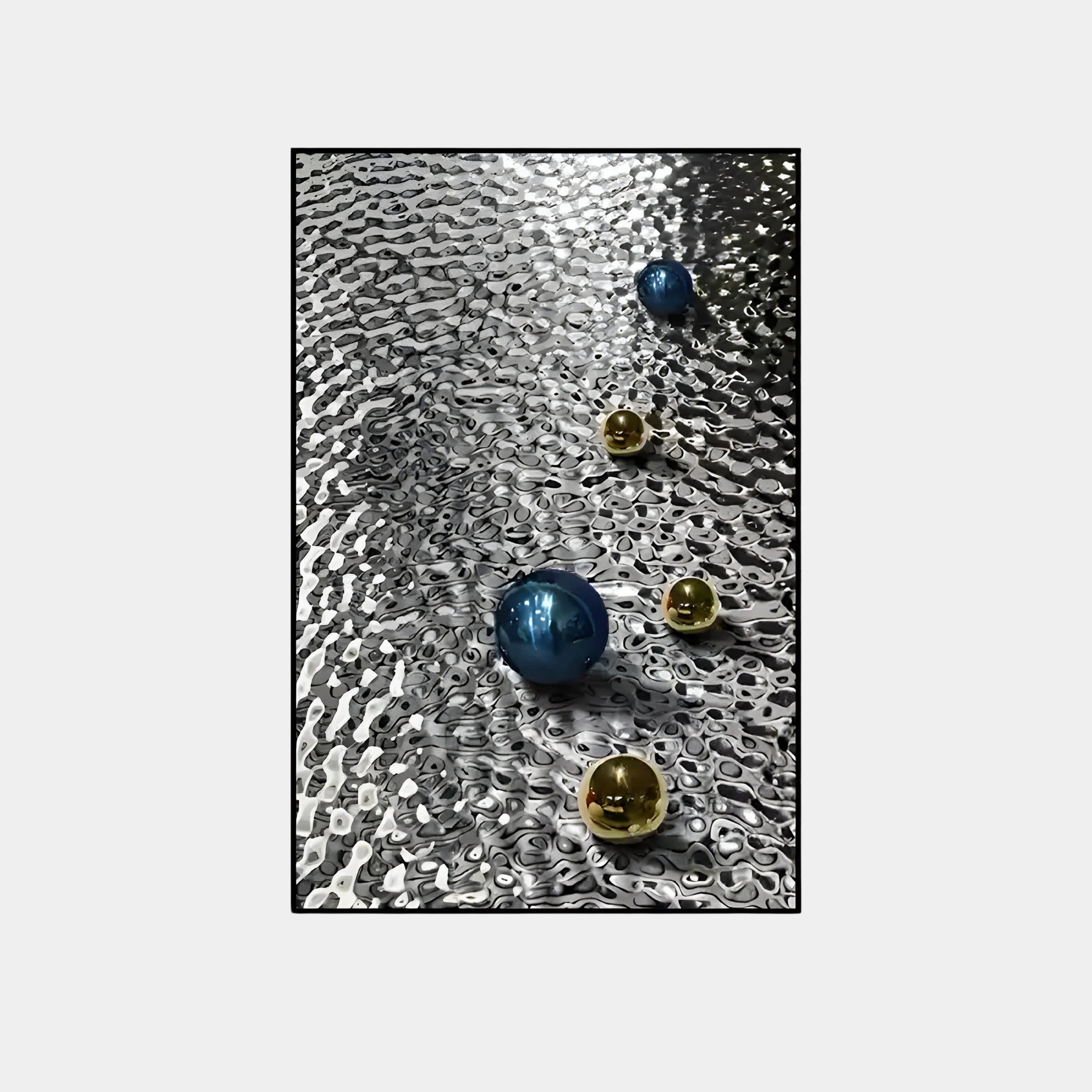 Four reflective Luminosa Blue & Gold Orbs by Giant Sculptures rest on textured metal, forming a diagonal line. Their contrasting colors and textures create rippled light patterns, enhancing the allure of this striking wall art.