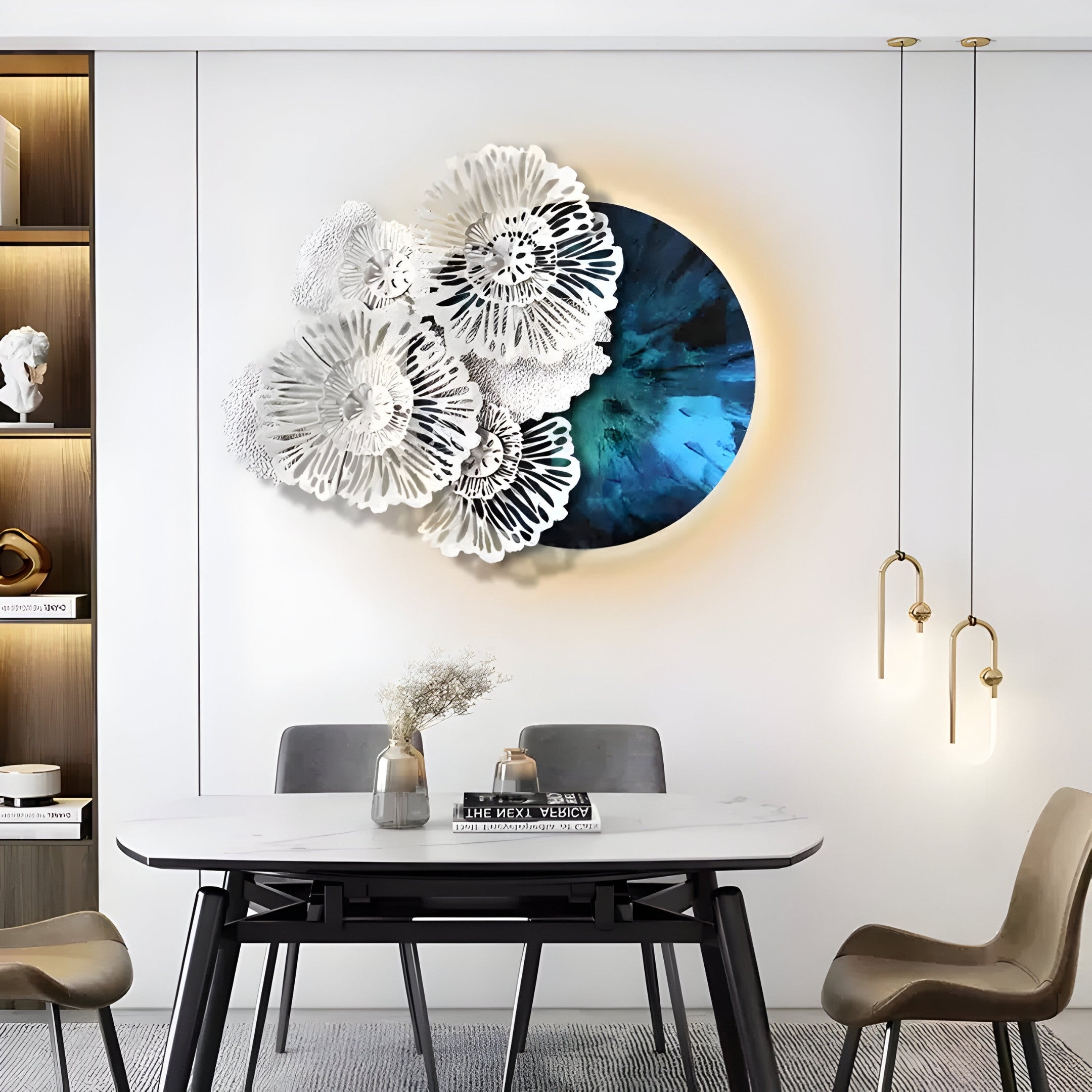 A modern dining area showcases a minimalist table and chairs, highlighted by the Giant Sculptures Aurum Flora White Petals Sapphire Metal Wall Art as its centerpiece. Two pendant lights hang overhead, while shelves displaying decor pieces enhance the contemporary vibe.