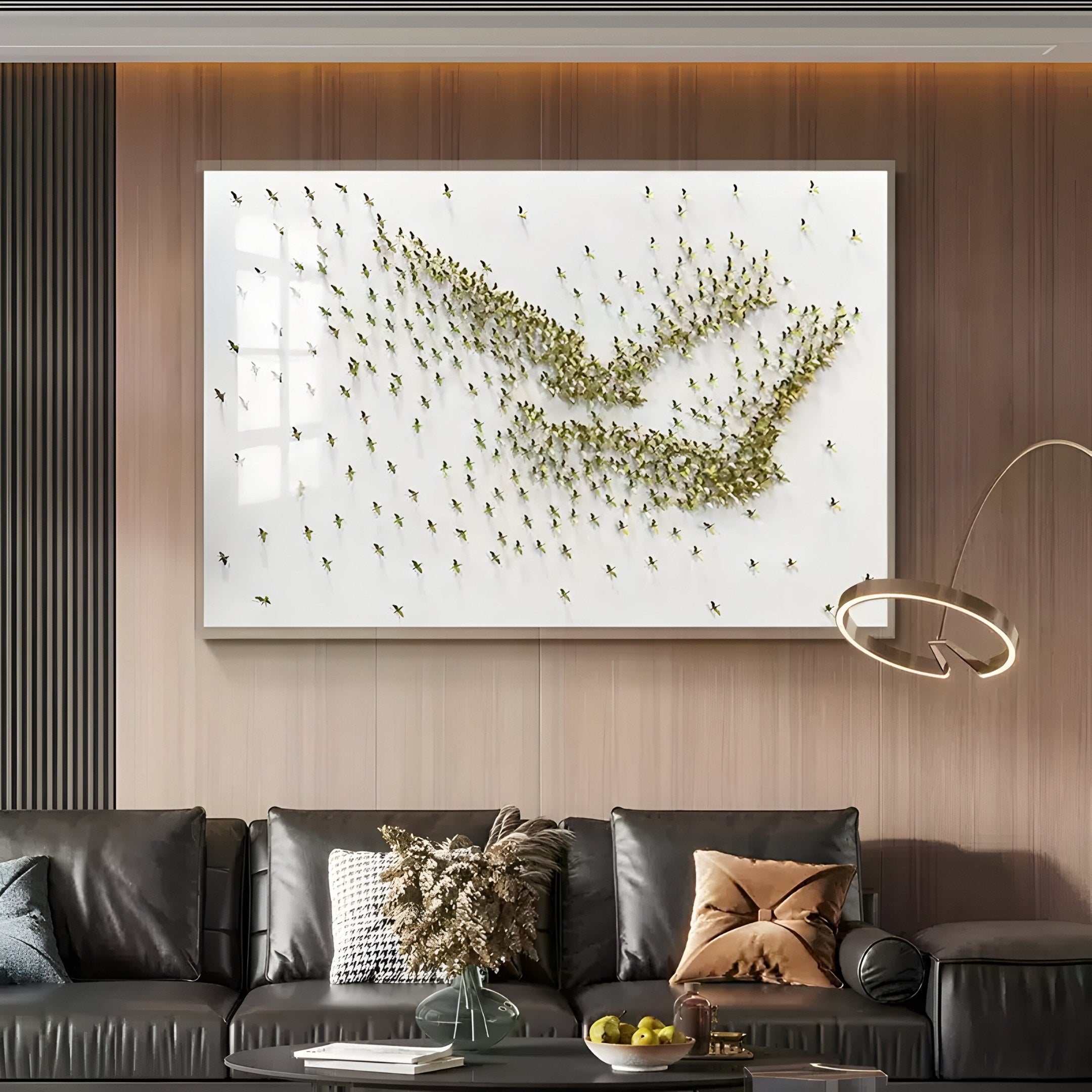 The Flock Golden Flight Sculpture Metal 3D Wall Art by Giant Sculptures adorns the wall above a black leather sofa. Minimalist aesthetics are enhanced by two cushions, a coffee table with dried flowers, and a modern lamp arching nearby, creating an elegant ambiance.