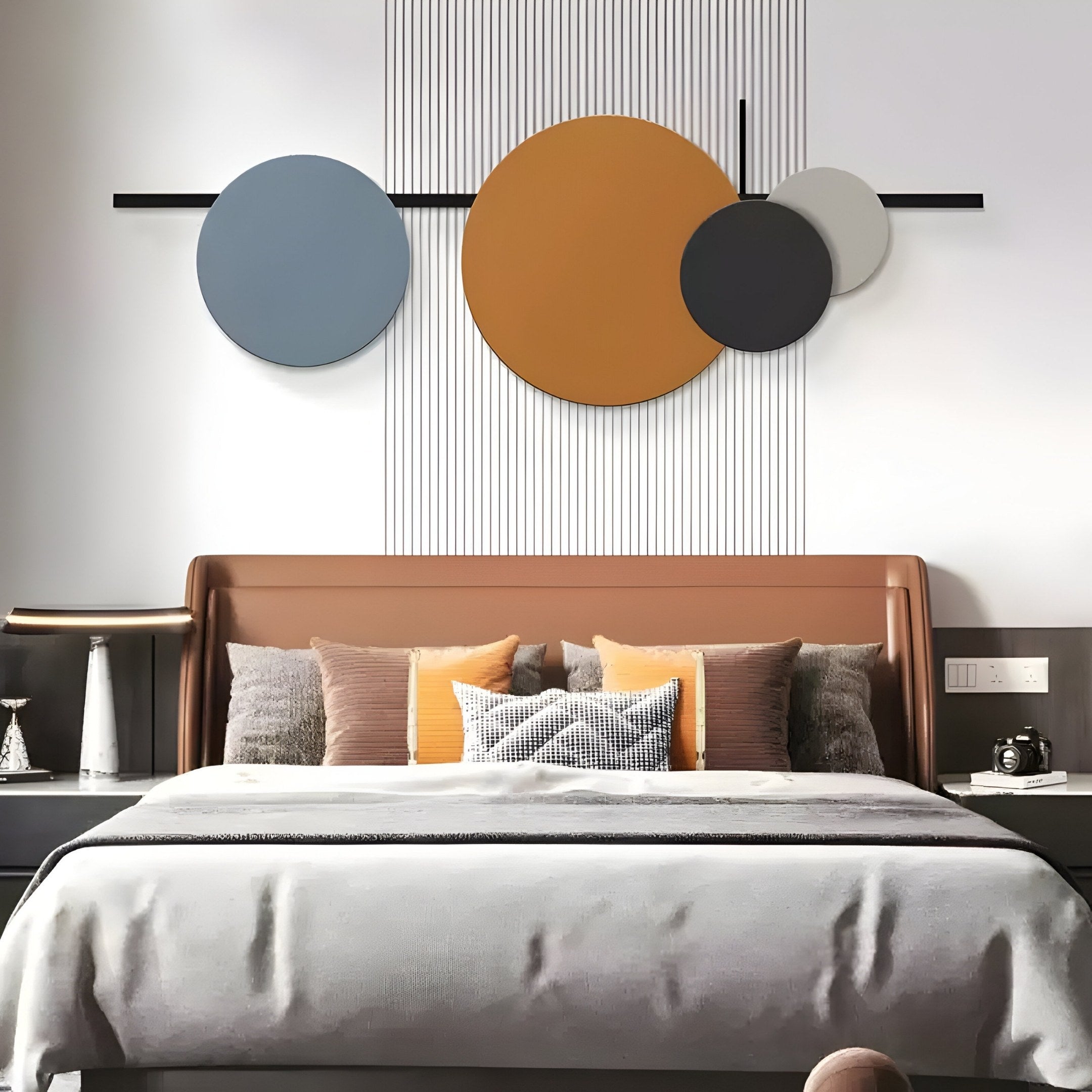 A modern bedroom showcases a brown headboard with gray and orange pillows on a neutral bedspread. Above, the Orbit III Dynamic Flow Multicoloured Circular Wall Art by Giant Sculptures adds geometric elegance with large blue, orange, black, and gray circles, enhancing the sleek interior design.