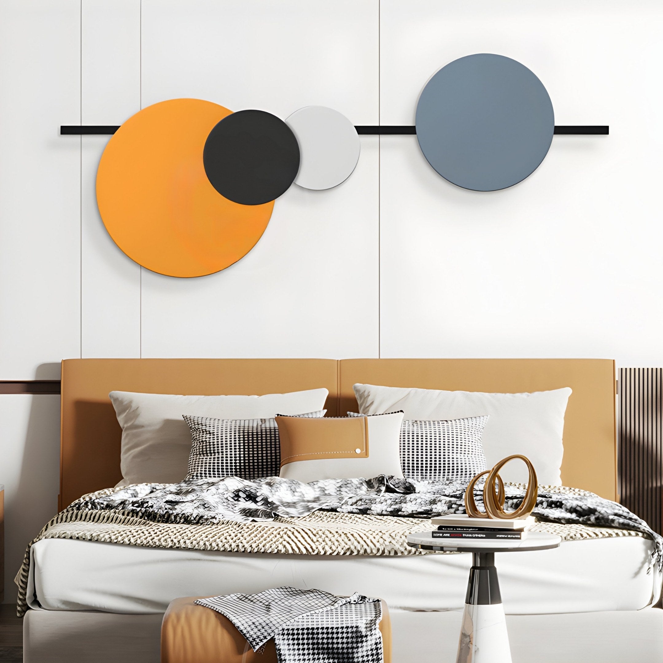 A modern bedroom features a beige bed with patterned pillows and a checkered throw. The setting is enhanced by Giant Sculptures Orbit III Radiant Balance Wall Art, displaying overlapping circles in orange, black, white, and gray. A small round table completes the cozy yet stylish look.