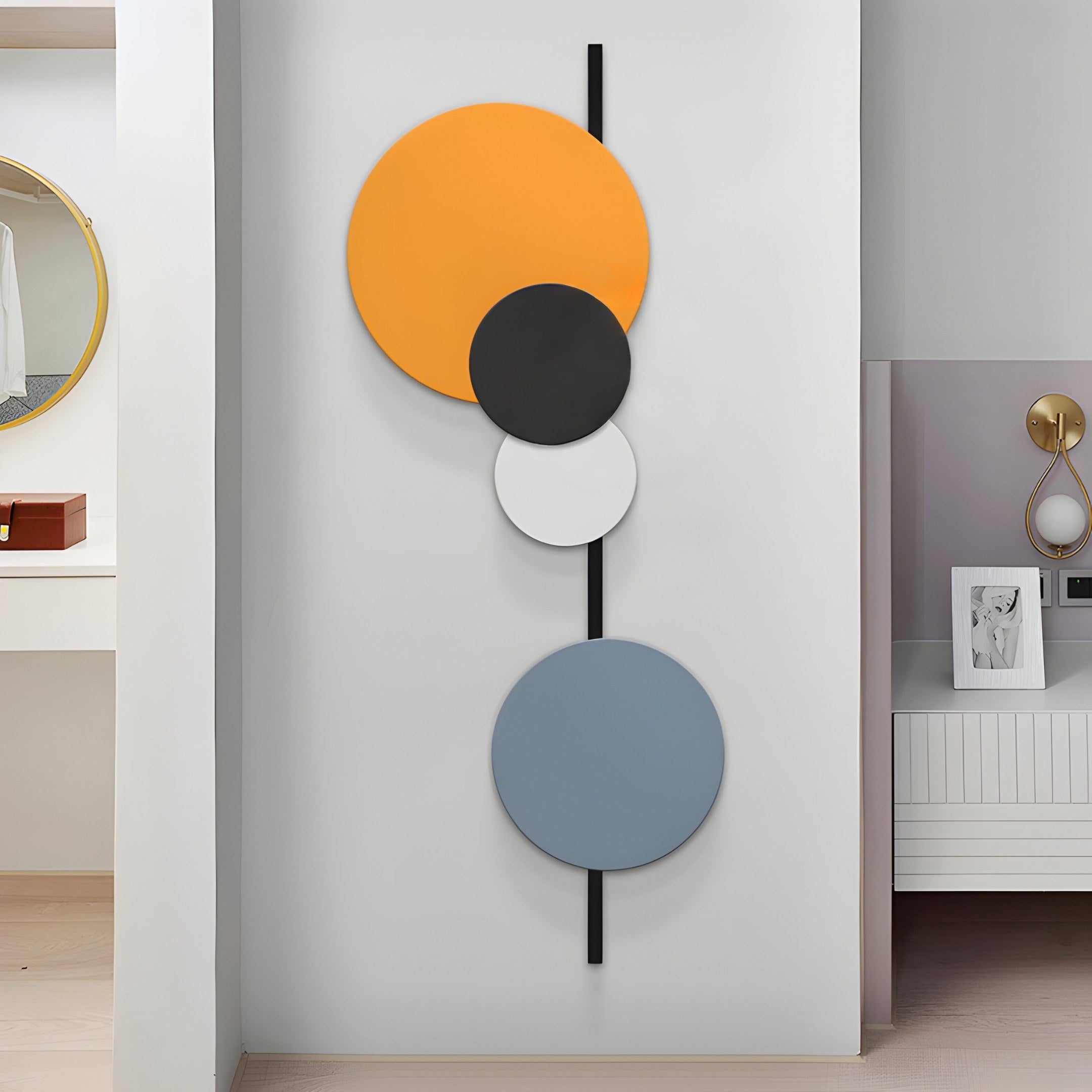 The Orbit III Radiant Balance Multicoloured Circular Wall Art by Giant Sculptures features four vertically aligned circles in orange, black, white, and blue on a light background. A nearby vanity and mirror are partially visible, adding geometric balance to the decor.