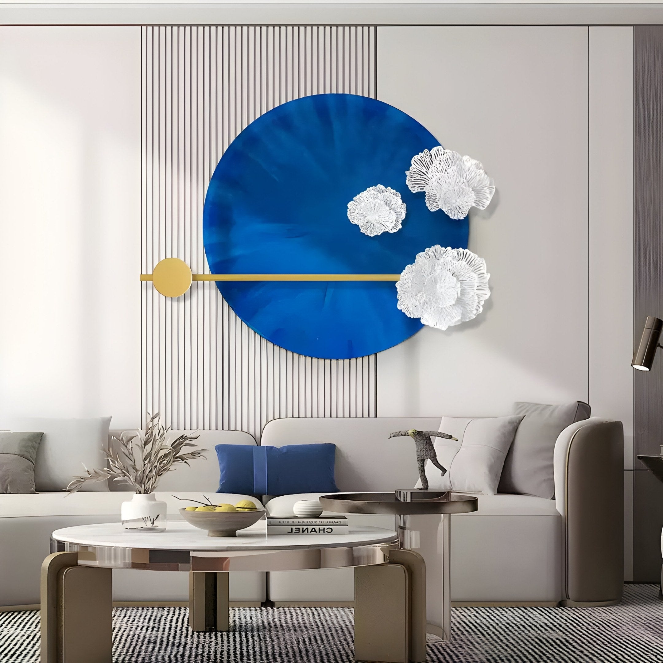 A modern living room features the Nova Floral Radiant Sapphire Blue Circular Wall Art from Giant Sculptures, showcasing white floral accents and a gold diagonal line. A gray sofa with pillows, a round coffee table with books and plants, and neutral decor epitomize refined modern interiors.