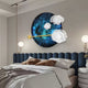 A modern bedroom features Giant Sculptures Nova Floral Radiant Azure Circular Wall Art above the bed, blending blue and white floral patterns. The dark blue upholstered bed is dressed with brown and white bedding, and two spherical pendant lights add a geometric touch on either side.