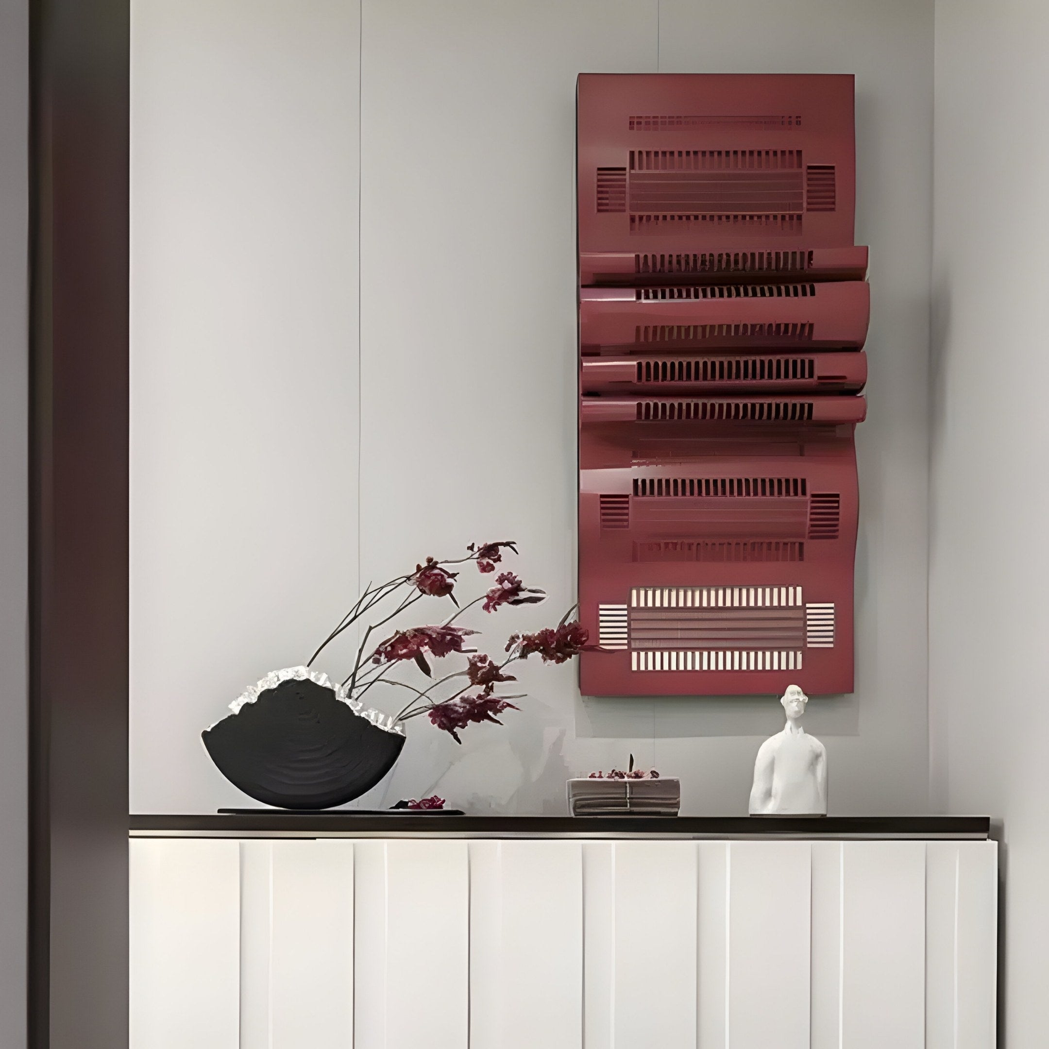 A minimalist interior features the Strata Red Vertical Folded Grid Metal Wall Art by Giant Sculptures above a white cabinet. A white figurine and black vase with red branches sit on top, while the soft gray wall enhances the bold, modern appeal in this contemporary space.