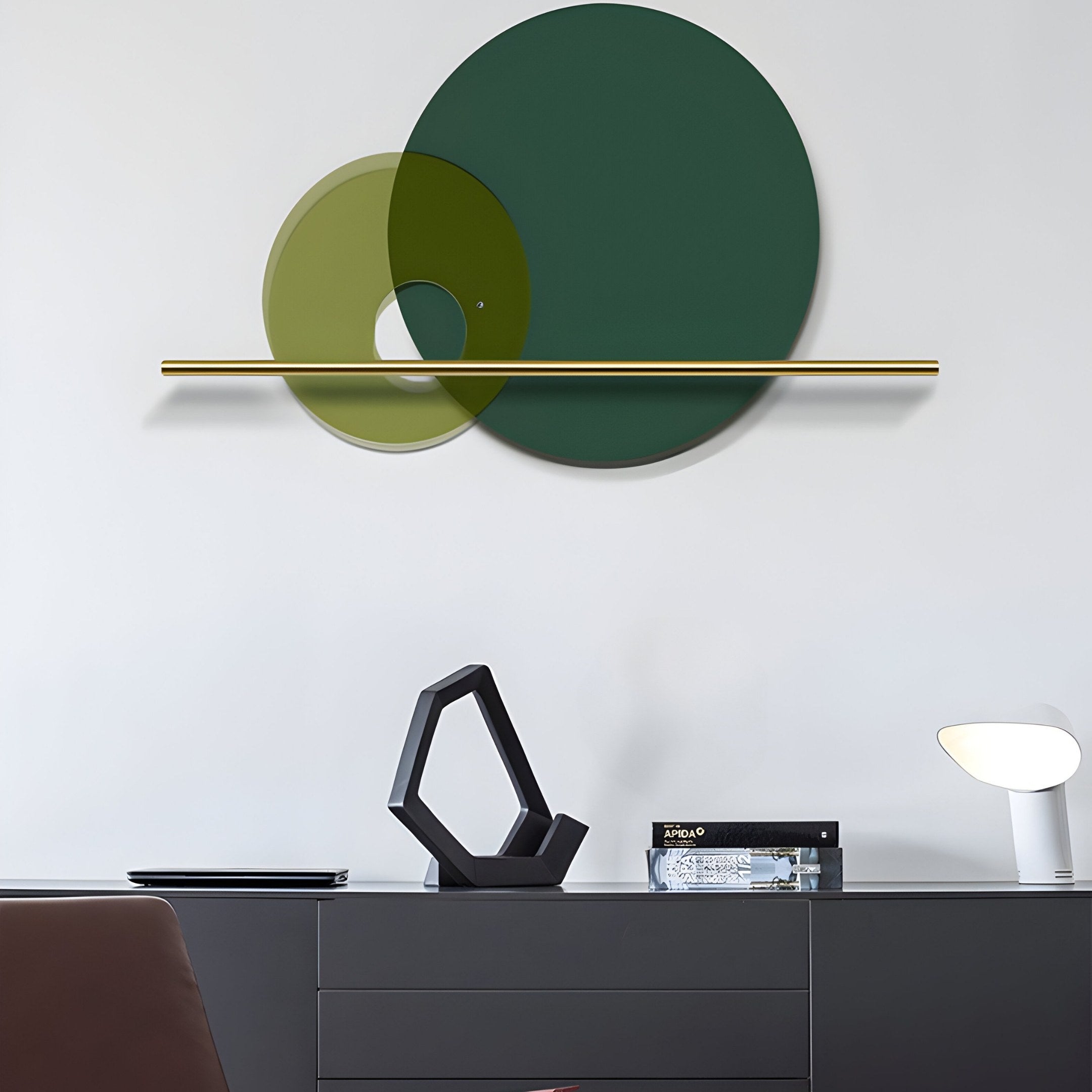 Giant Sculptures’ Luna Forma Forest Green Round Brass Accent Wall Art, featuring intersecting green and yellow translucent circles and a thin gold bar, complements a sleek black cabinet with geometric decor and a modern white lamp, enhanced by subtle brass accents for elegant harmony.