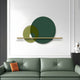 A stylish living room showcases a forest green leather couch accented with an orange cushion. Above it is the Luna Forma Forest Green Round Brass Accent Wall Art from Giant Sculptures, featuring concentric green and yellow circles intersected by a gold line. A white bust rests on a dark pedestal nearby.