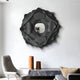 A modern living room showcases the Sculptura Mirror Charcoal Grey Floral Round Metal Wall Art by Giant Sculptures above a gray stone bench with two terracotta vases. A textured armchair to the left complements the scene, while a beige cushion on the bench adds elegance and cohesion.