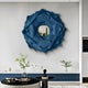 A modern living room features the Sculptura Mirror Midnight Blue Floral Round Metal Wall Art by Giant Sculptures. Beneath, a minimalist table displays a plant, small black sculpture, and another decorative object, creating an elegant ambiance with neutral tones and contemporary decor.