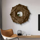 A modern interior features the Sculptura Mirror Sand Beige Floral Round Metal Wall Art by Giant Sculptures, below which a minimalist shelf with books and a glass device sits. A chair with a sand beige leather seat is partially visible next to a gray curtain.