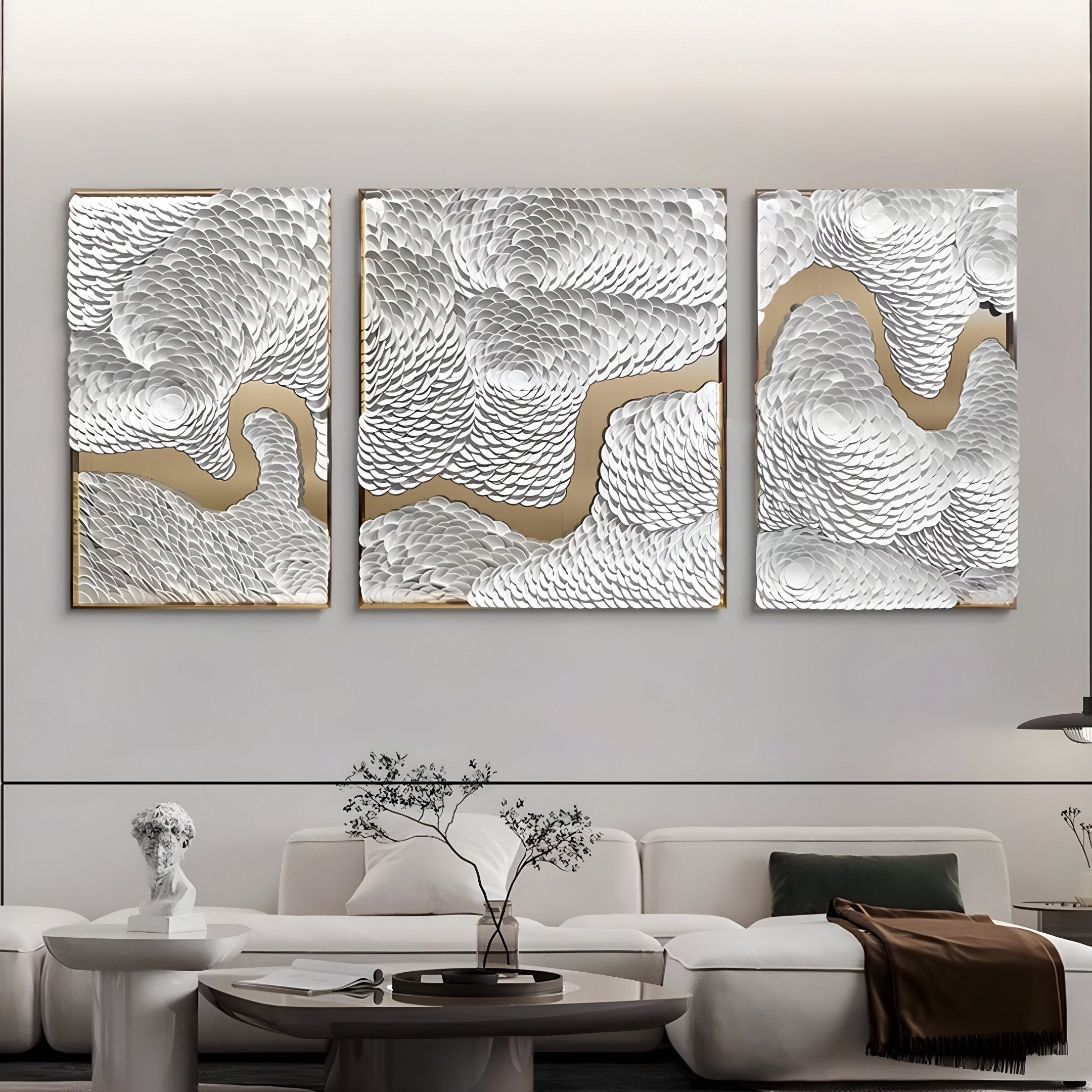 A modern living room showcases a pearl white textured sofa with a brown blanket and a round coffee table, adorned with a potted plant. The highlight is the Riverine Flow Pearl White Textured Triptych Wall Art by Giant Sculptures, featuring swirling textures and gold accents above the sofa.