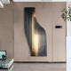 On a beige wall, the Giant Sculptures Celestial Gold Radiance Curved Angled Abstract Metal Wall Art showcases vertical gold lines on a dark panel. A small plant and control panel complement the minimalist rooms polished floor and a small black device on a ledge.