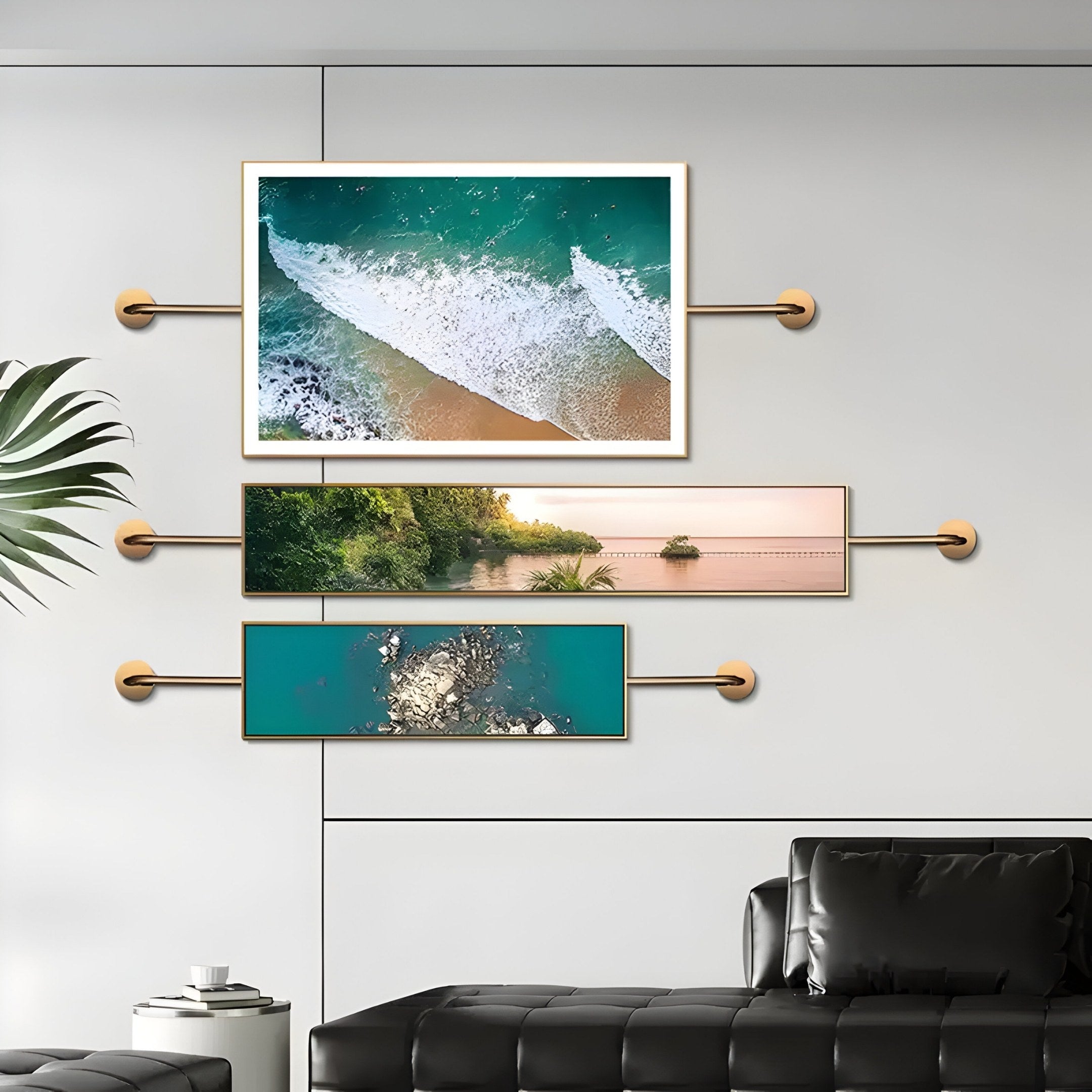 Three pieces of Giant Sculptures Visario Ocean & Nature Inspired Metal Wall Art, featuring ocean waves, a coastline, and aerial sea views with rocks in gold frames, are displayed on a minimalist wall above a leather sofa. A potted plant enhances the coastal theme.