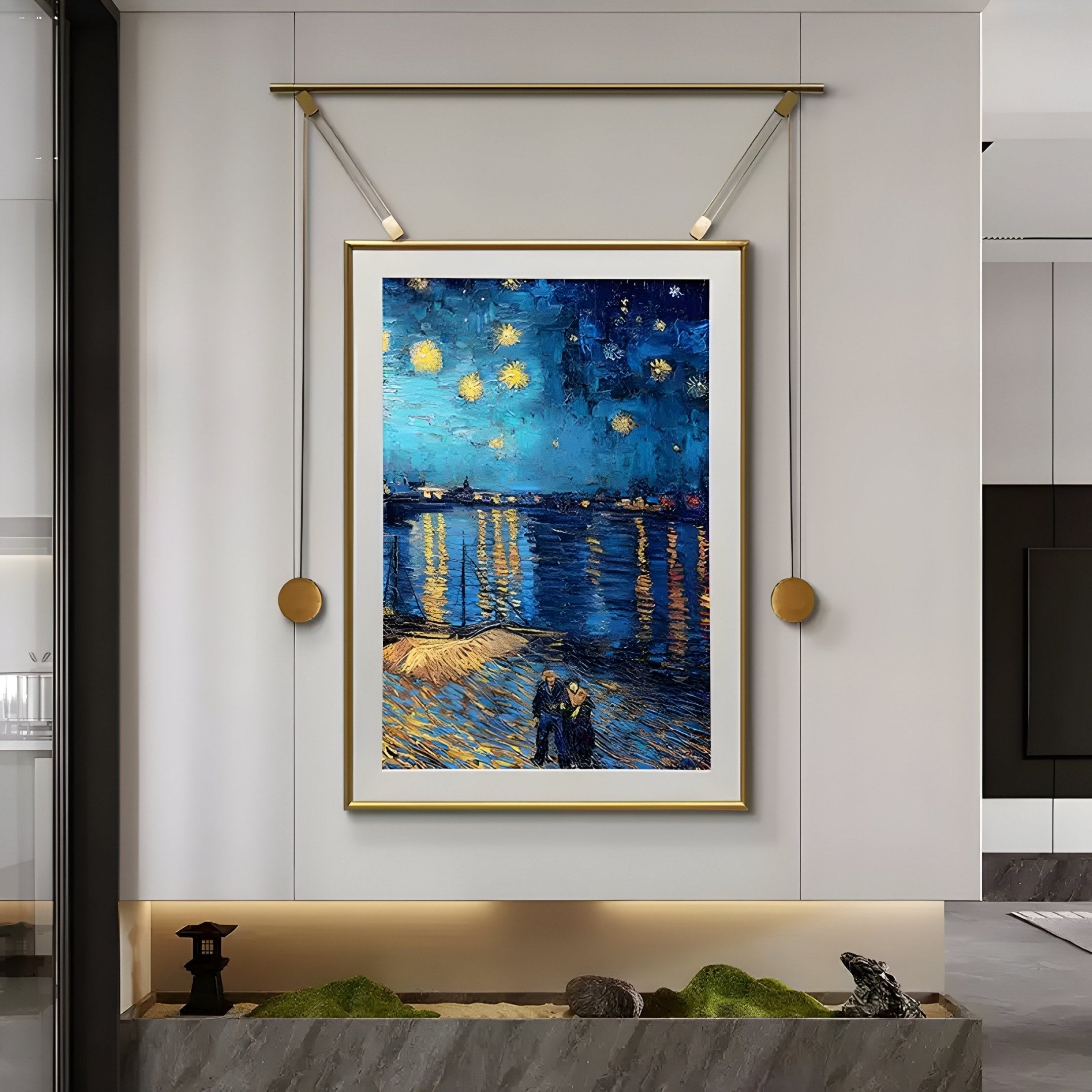 The Reverra Starry Night Over The Rhone Hanging Frame Wall Art by Giant Sculptures, featuring a couple strolling along a starry shoreline, is elegantly displayed on a white wall. The gold-framed piece enhances the room’s minimal decor with its plants and stone accents.