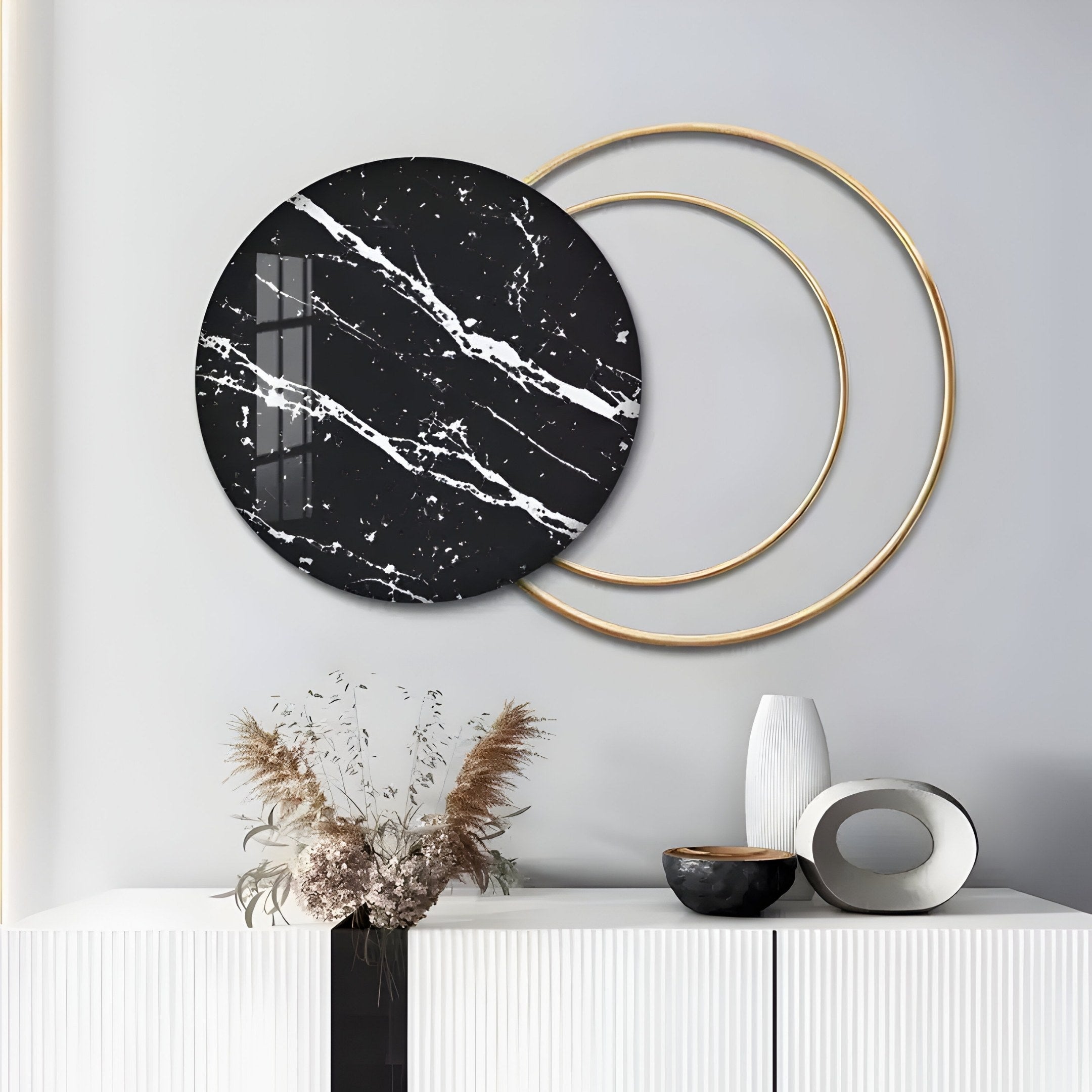 In a modern interior, a white cabinet is adorned with ceramics and pampas grass. Above it, the Eclipse Luxe Black Noir Marble Circular Ring Wall Art by Giant Sculptures features a black marble circle layered with two gold rings on a gray wall.