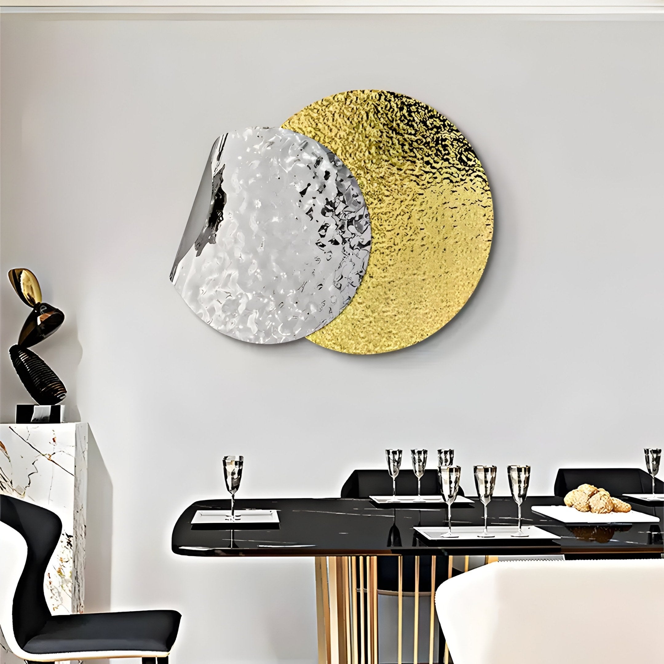 A modern dining room boasts a black table adorned with glassware and monochrome chairs. Contemporary art enhances the wall, featuring Giant Sculptures Metaluxe II Gold & Silver Dual Disc Textured 3D Wall Art. In the corner, another sculpture rests on a sleek marble surface.