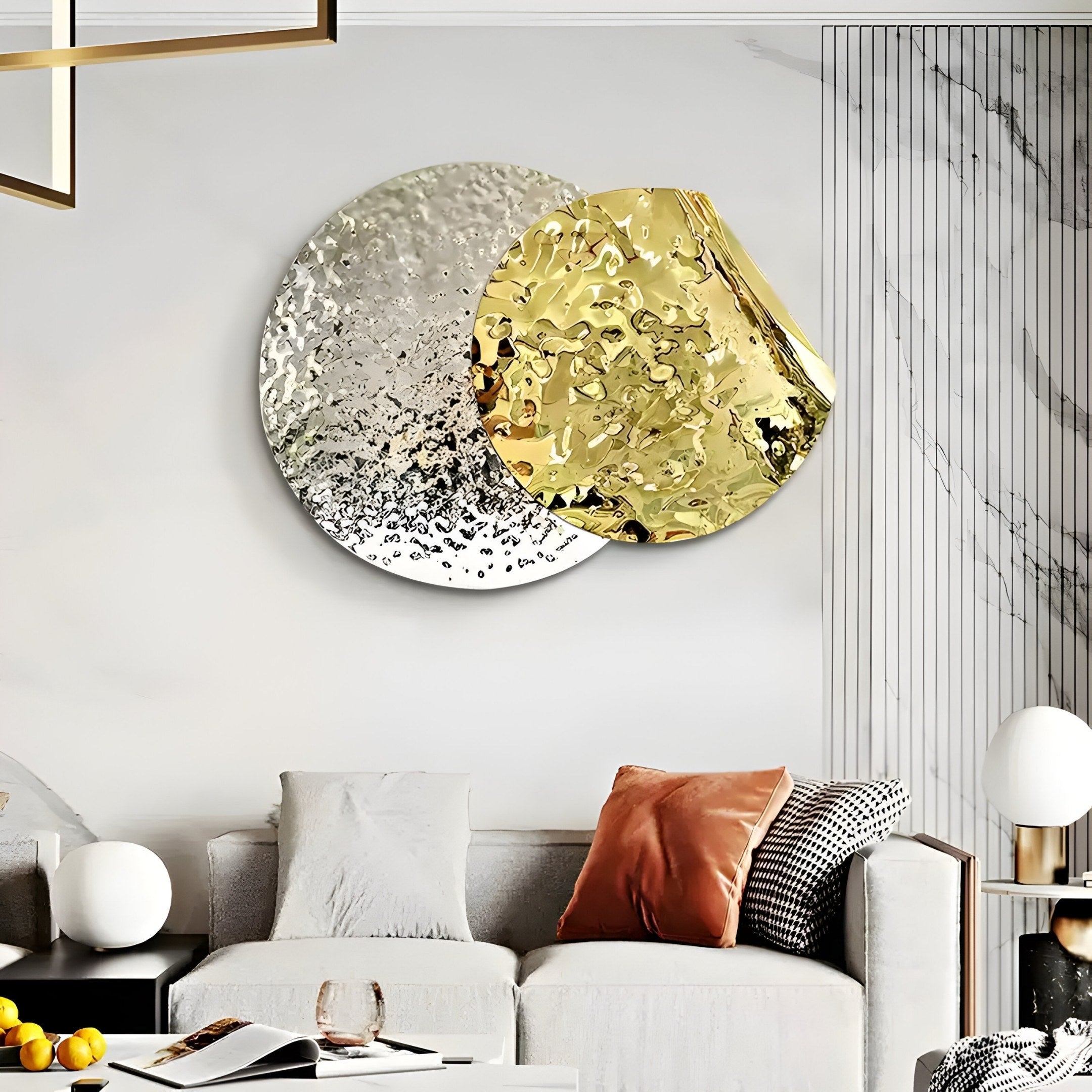 A modern living room features a white sofa with gray, black, and orange cushions. The space is enlivened by Giant Sculptures Metaluxe III Gold & Silver Dual Disc Textured 3D Wall Art. A side table with a white lamp and fruit bowl finishes the decor.