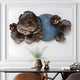 Displayed above the table is the Luminis Bloom Blue Floral Metal Wall Art by Giant Sculptures, featuring hand-crafted flowers with a central blue circle. The table holds a vase with greenery and a decorative bear-like object, surrounded by chairs in this modern setting.