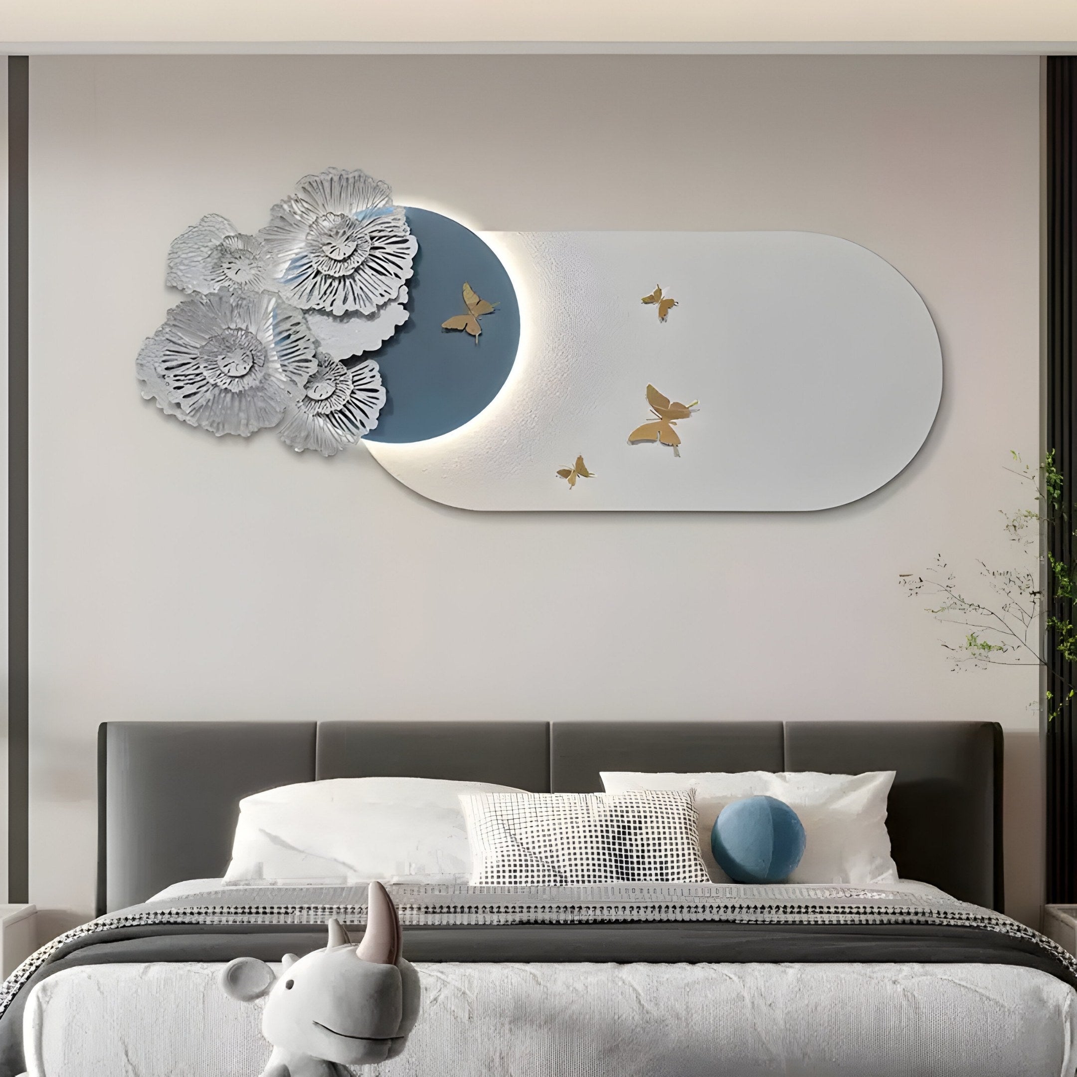 A modern bedroom showcases sleek decor, featuring the Flutter Bloom Blue Butterfly Floral 3D Wall Art by Giant Sculptures above the bed. This piece displays silver flowers and golden butterflies on a white and blue circle. The bed includes white bedding, a gray headboard, and a decorative gray cushion.