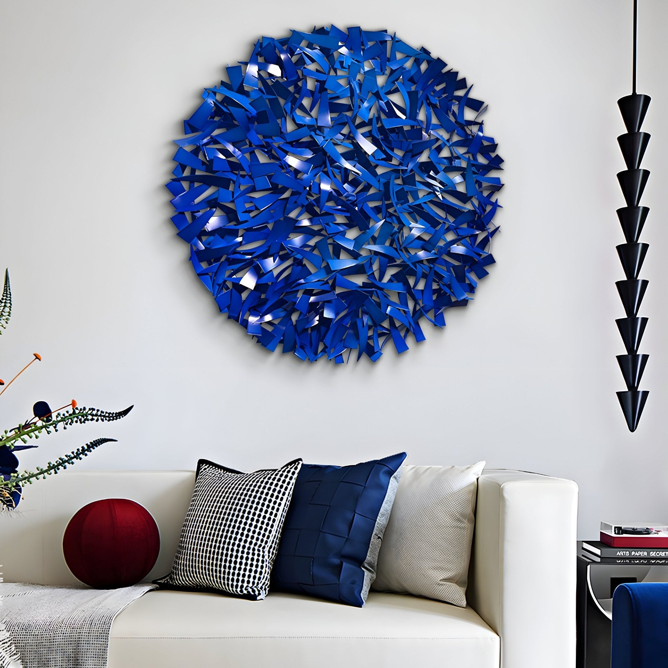 A modern living room showcases a white couch with black, white, and cobalt blue cushions. Above it hangs the Eclat Sphere Blue Abstract Metal Wall Art by Giant Sculptures. To the right is a black cone pendant light, while plants subtly appear from the left.