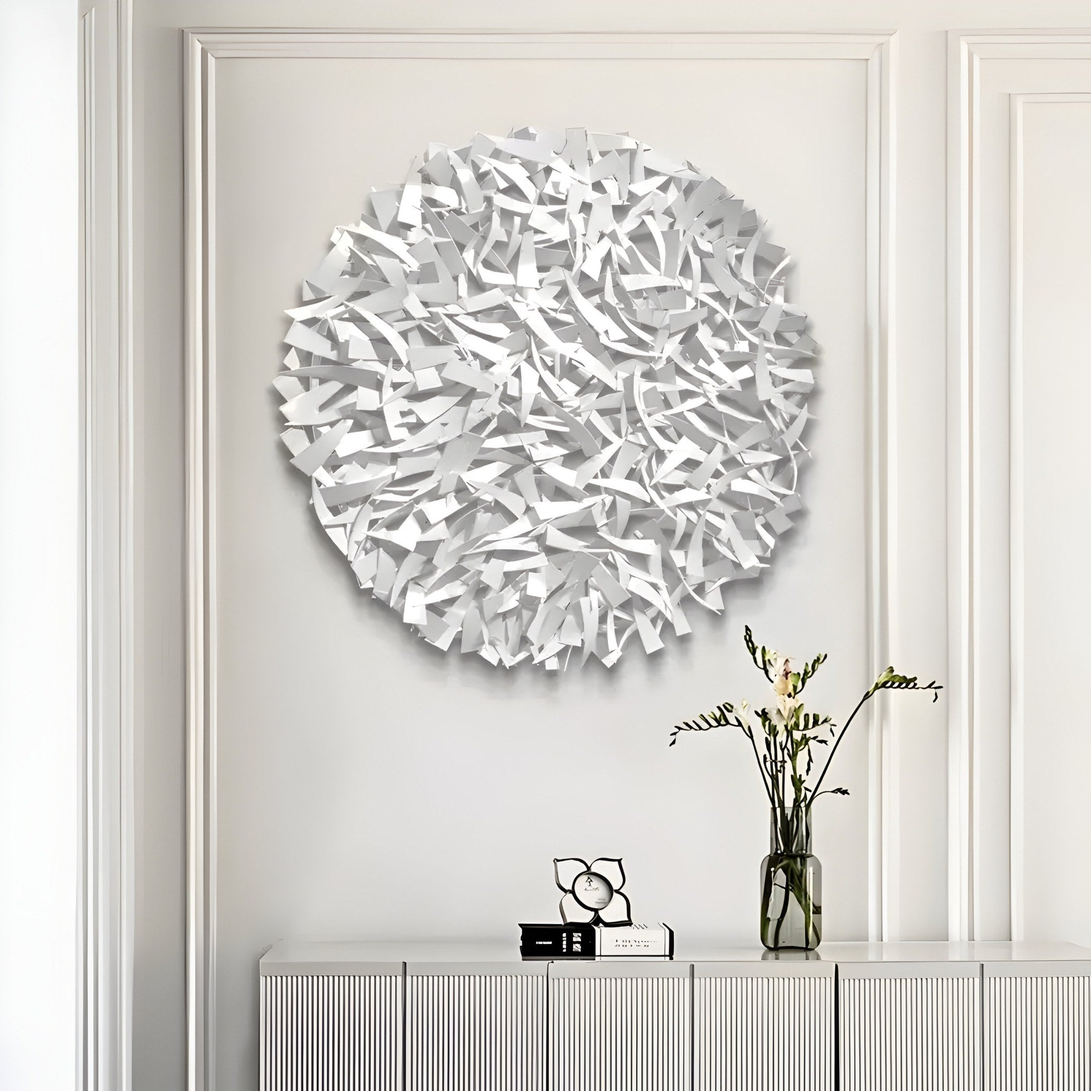 The Eclat Sphere White Abstract Metal Wall Art by Giant Sculptures is mounted above a minimalist white sideboard. A tall clear vase with delicate green and white flowers and a small abstract clock complete this captivating display.