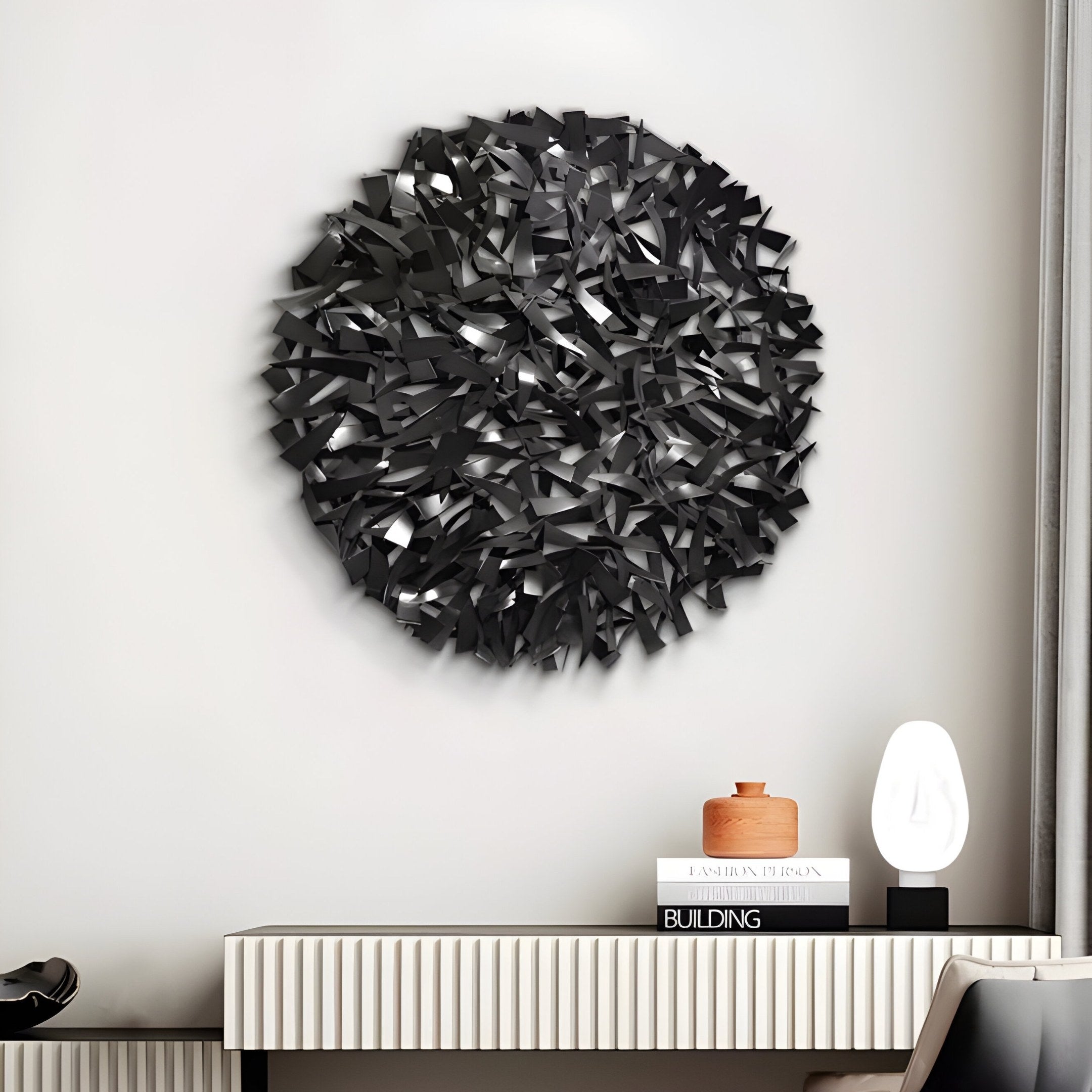 A modern living room features the Eclat Sphere Black Abstract Metal Wall Art by Giant Sculptures above a console table. The table holds a book, a terracotta pot, and an illuminated white lamp, all set against a neutral-hued wall.