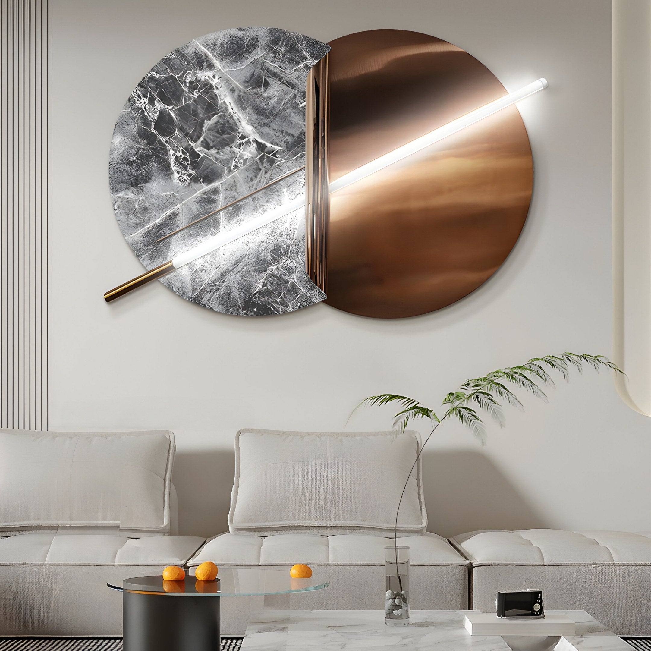 The modern living room showcases Giant Sculptures Lunaris Grey Marble & Bronze LED Wall Art, featuring two circular panels connected by a diagonal light bar. It also includes a cream sofa, cushions, wiry plant, and marble coffee table with three oranges.