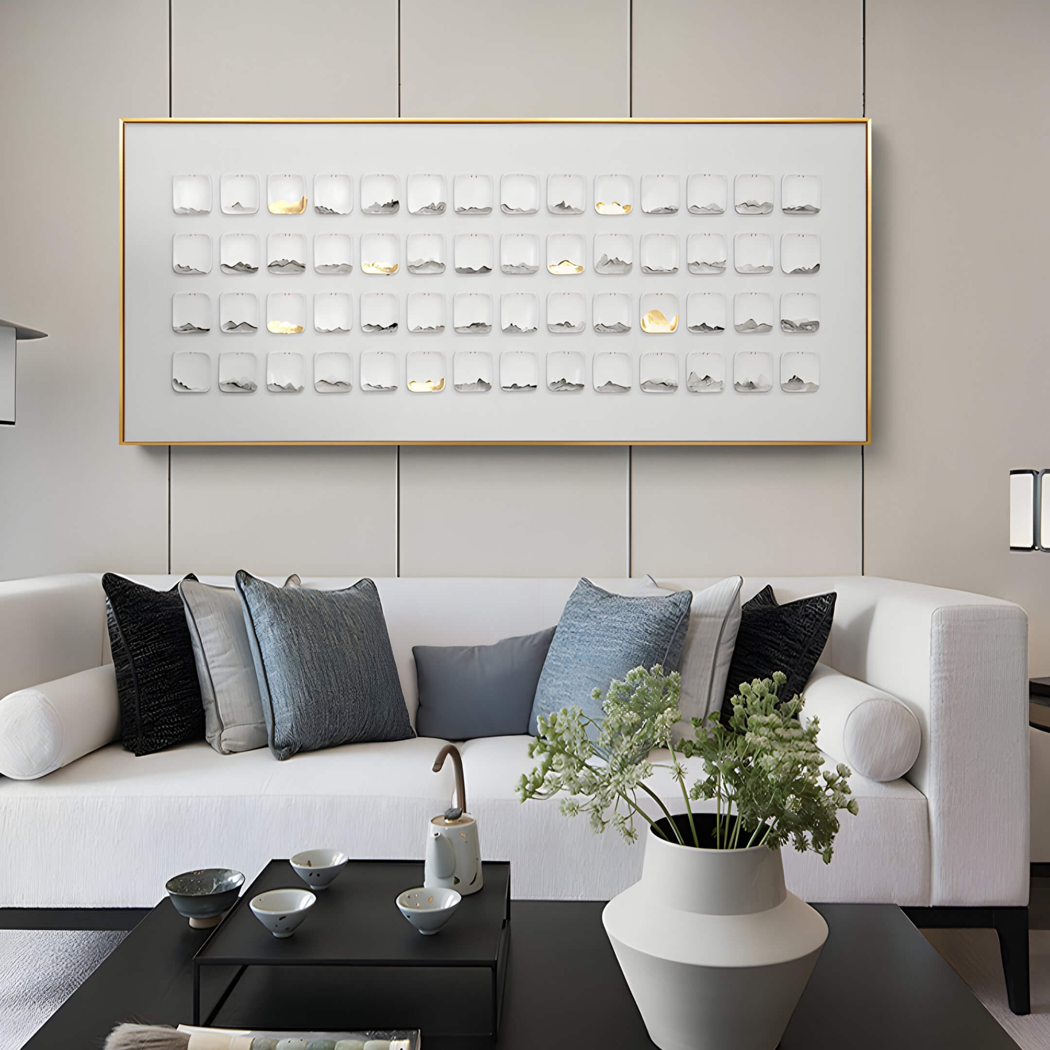 A modern living room features a white sofa with blue and gray pillows, a coffee table displaying a decorative tray and vase. Above the sofa hangs the Lureva 3D Ceramic Wall Installation With Gold Foil & Metal Trim by Giant Sculptures, showcasing abstract square elements.