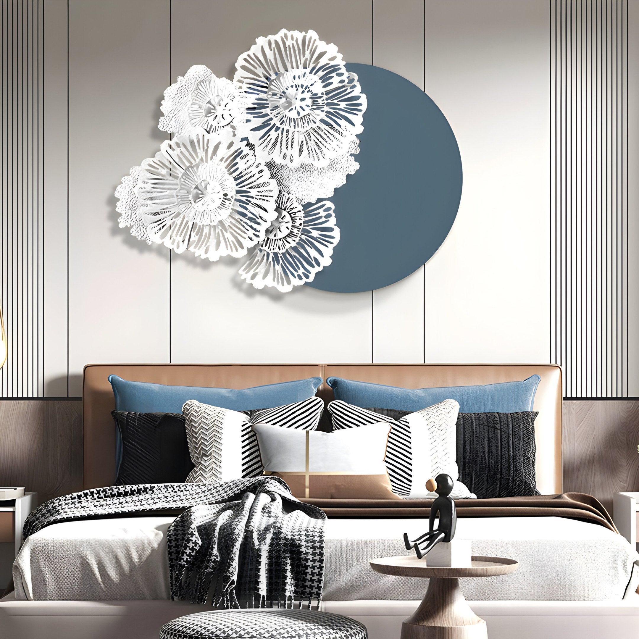 A contemporary bedroom showcases a beige leather bed with blue and black pillows, a black and white patterned throw, and a figurine on a round table. The highlight is the Fleur Halo Grey Round White Metal Floral Decorative Wall Art by Giant Sculptures, featuring white flowers over a large blue circle.