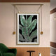 The Glacia Green Botanical Crystal Hanging Framed Wall Art by Giant Sculptures features green botanical illustrations with white outlines on a black background, accented with golden fixtures. It complements a modern interior including a green chair and wooden side table, epitomizing contemporary design.