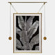 The Glacia Monochrome Botanical Crystal Hanging Framed Wall Art by Giant Sculptures features detailed white banana leaves on black, set in a gold frame with rods and golden weights, showcasing contemporary craftsmanship.