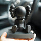 A person holds a Midnight Black I Iconify Bobblehead Figure - 10cm by Giant Sculptures, featuring large, rounded features and spiral-like head shapes. It has a modern design on a square base against a blurred steering wheel backdrop.