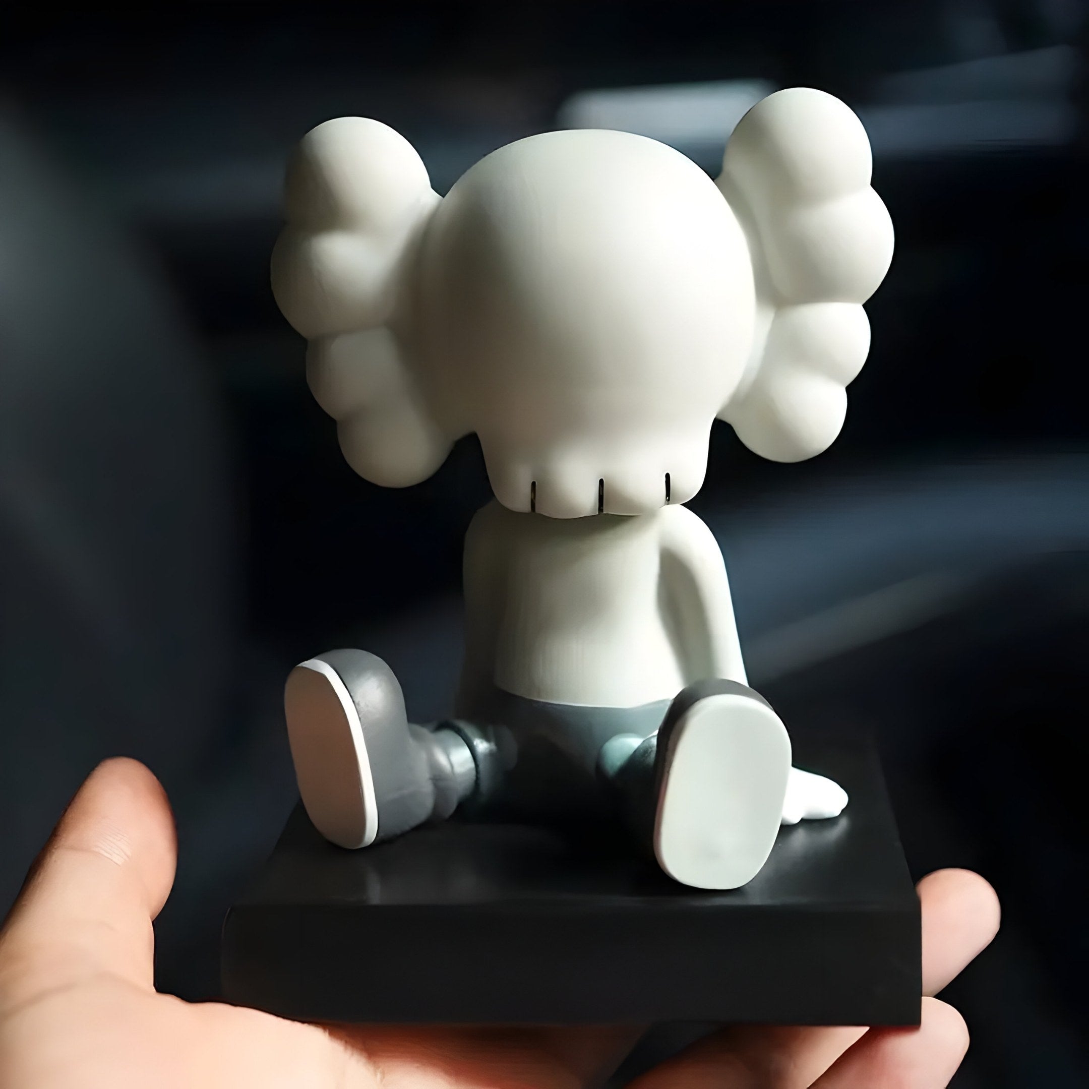 A hand holds the Dove Grey Iconify Bobblehead Figure by Giant Sculptures, featuring a cartoonish design with a bulbous head and cloud-shaped ears. This 10cm resin collectible elegantly sits on a black base, showcasing minimalist aesthetics with its back to the viewer.