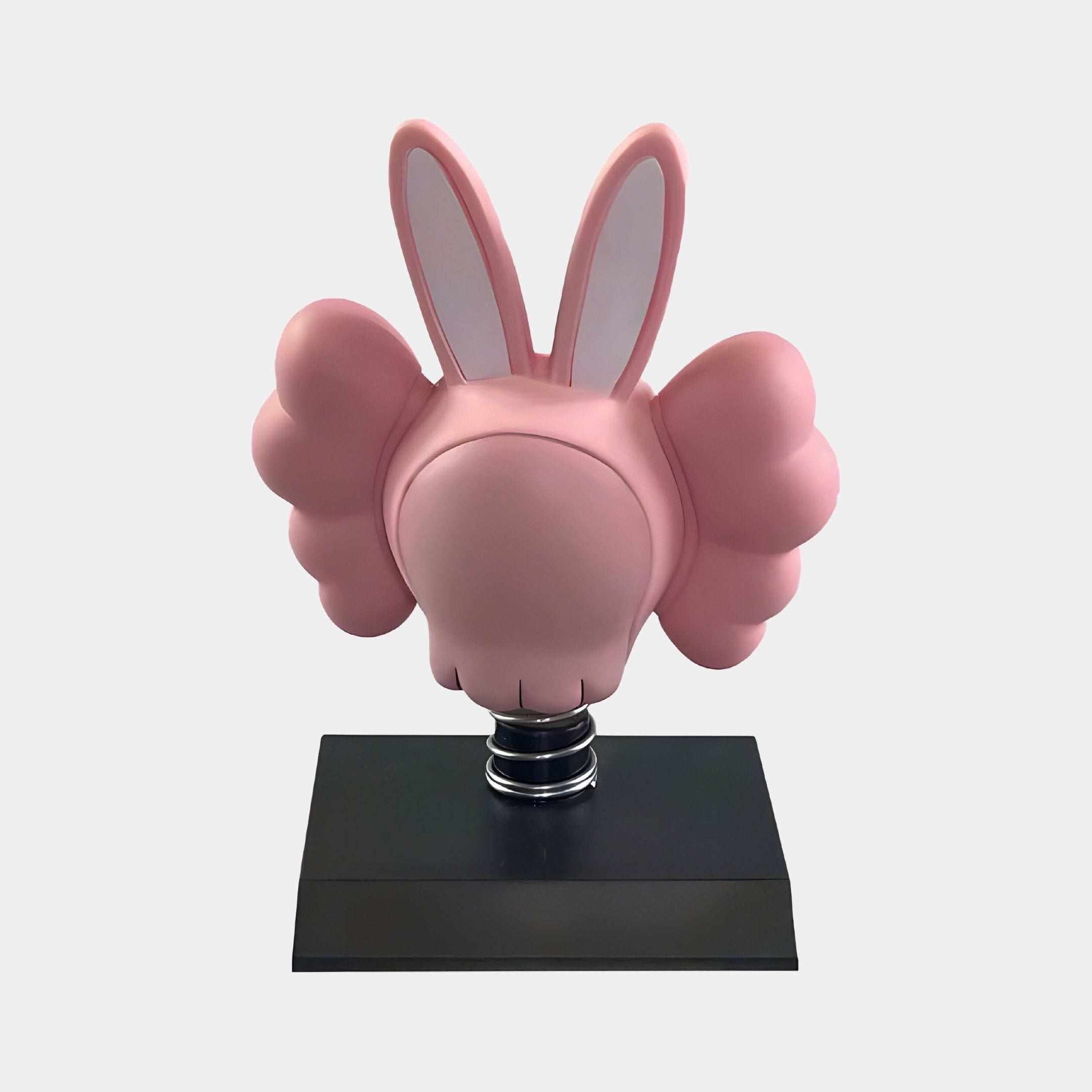 The Petal Pink Iconify Bobblehead Rabbit Figure by Giant Sculptures features large bunny ears and bulbous wings. It sits on a black rectangular base with a featureless, spherical head supported by a coiled spring, reminiscent of a bobblehead rabbit. Total height is 10cm.