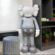 A 100cm Soft Grey Iconify Standing Sculpture from Giant Sculptures, featuring cartoon-like design with crossed-out eyes, rounded features, and glove-shoe details, stands in a modern room. Its fluffy head shapes complement a gray stool, shelf, and potted plant for a minimalist elegant look.