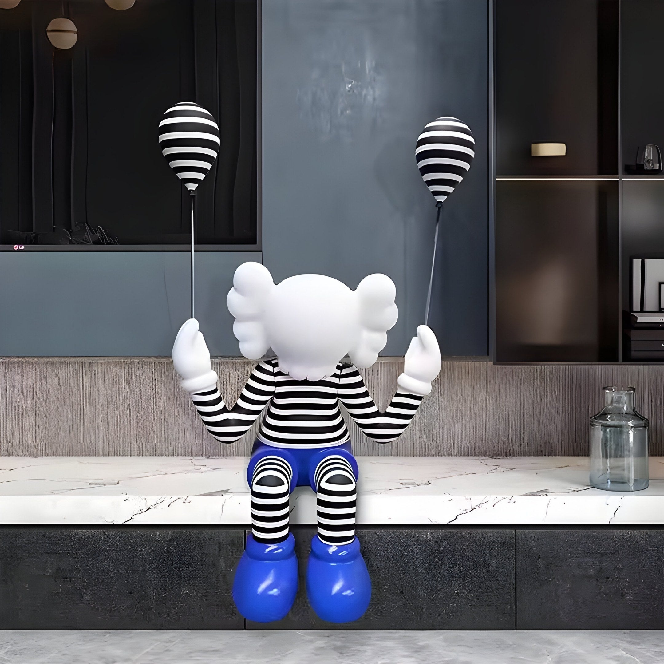 The Deep Blue Iconify Mime Sitting Sculpture by Giant Sculptures, at 195cm, showcases a luxurious seated figure with a cartoonish head in black and white stripes and blue shoes, holding two striped balloons, set against sleek shelves on a gray wall.